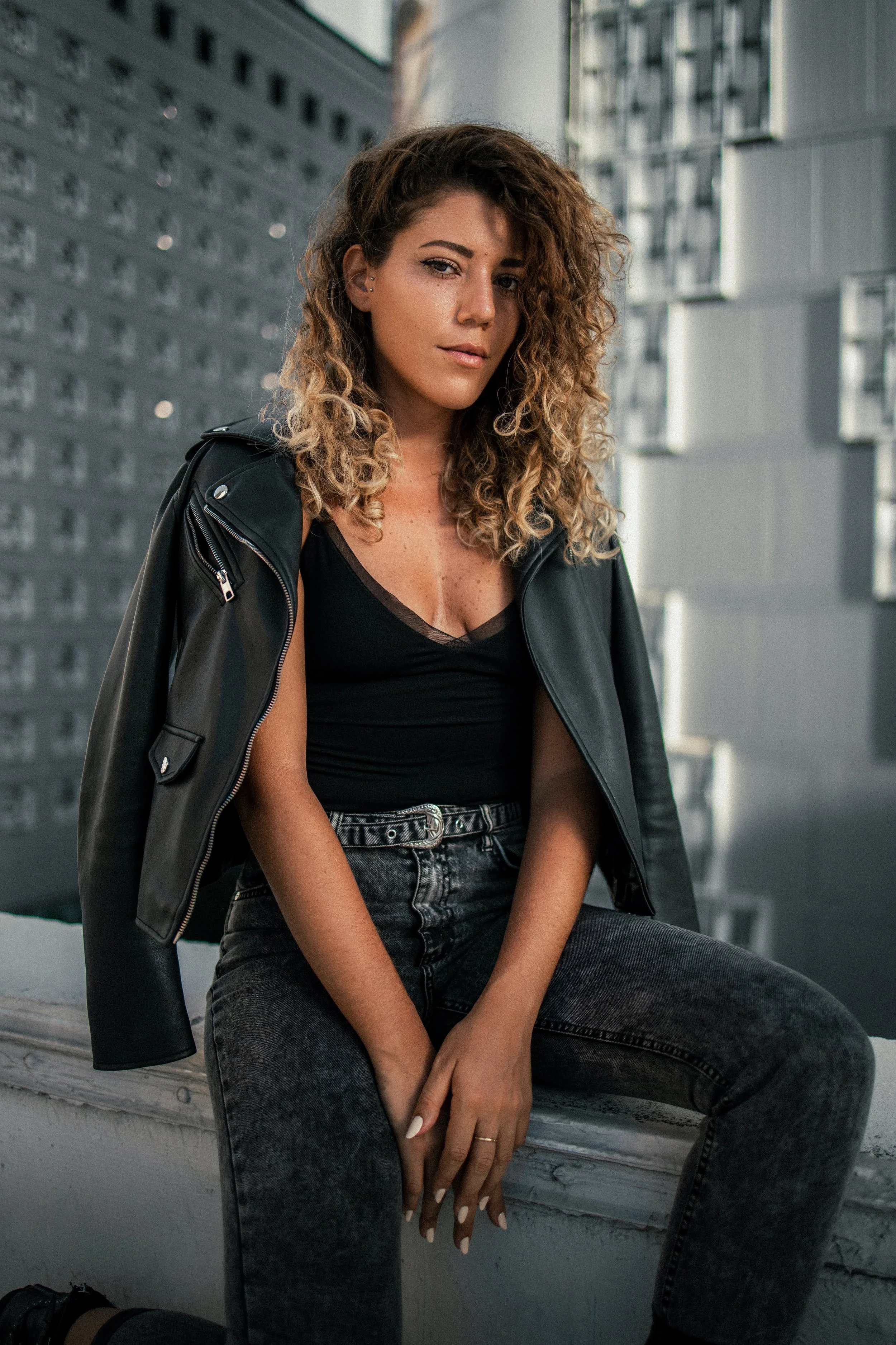 A young woman with curly brown hair, wearing a black top, black leather jacket, and black jeans, sitting on a concrete ledge in an urban setting with modern apartment buildings in the background.
