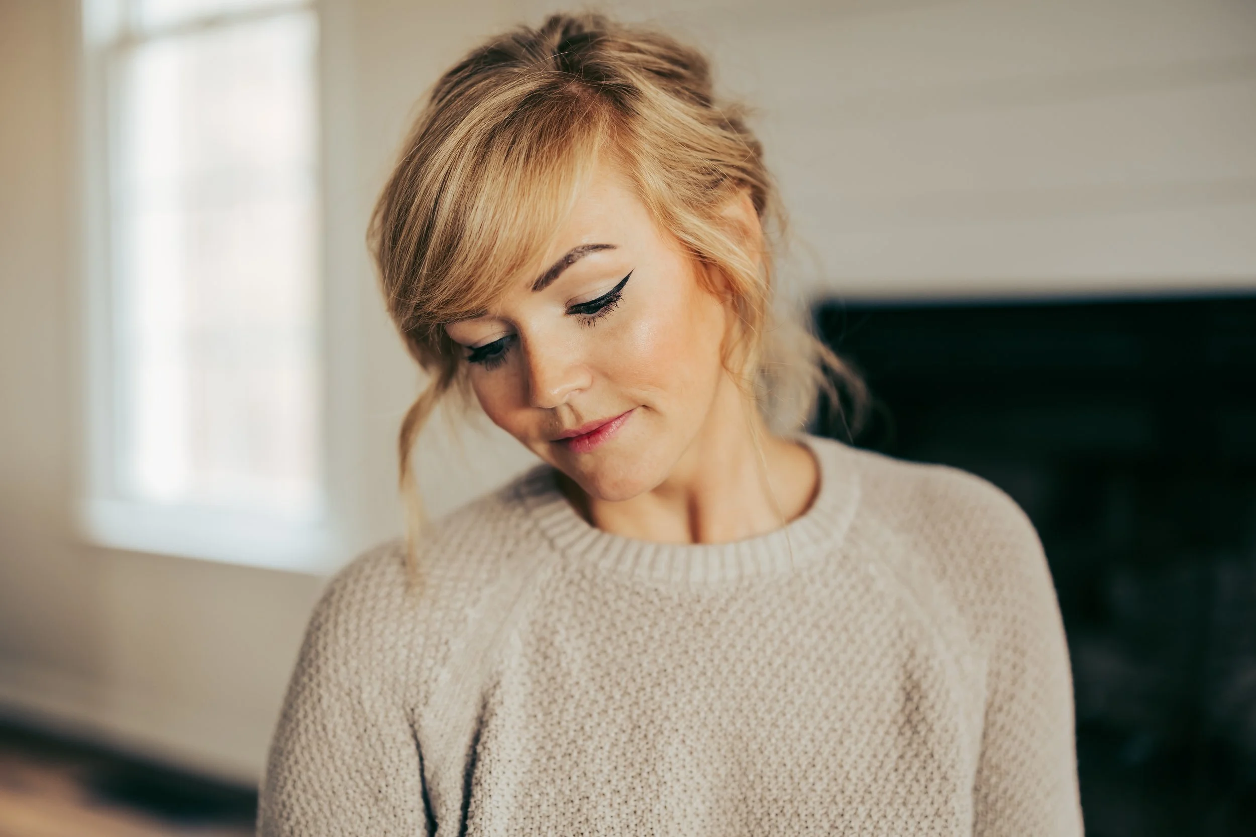 A woman with blonde hair and winged eyeliner looking down in a cozy indoor setting.