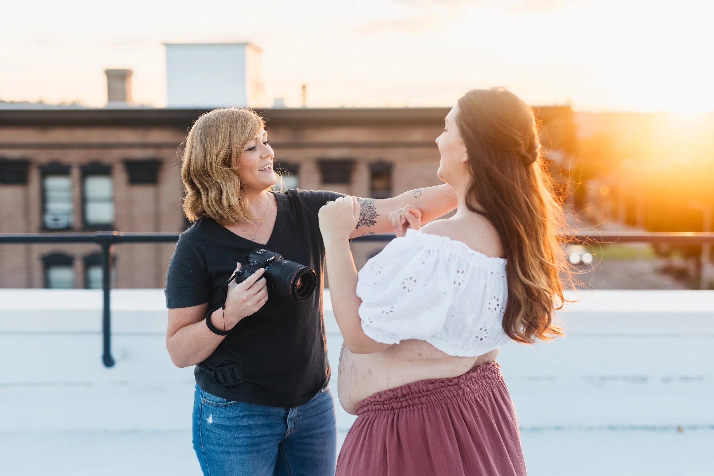 Two women on a rooftop at sunset, one with a camera, smiling and touching the other's arm, the other wearing a white off-shoulder top and burgundy skirt.
