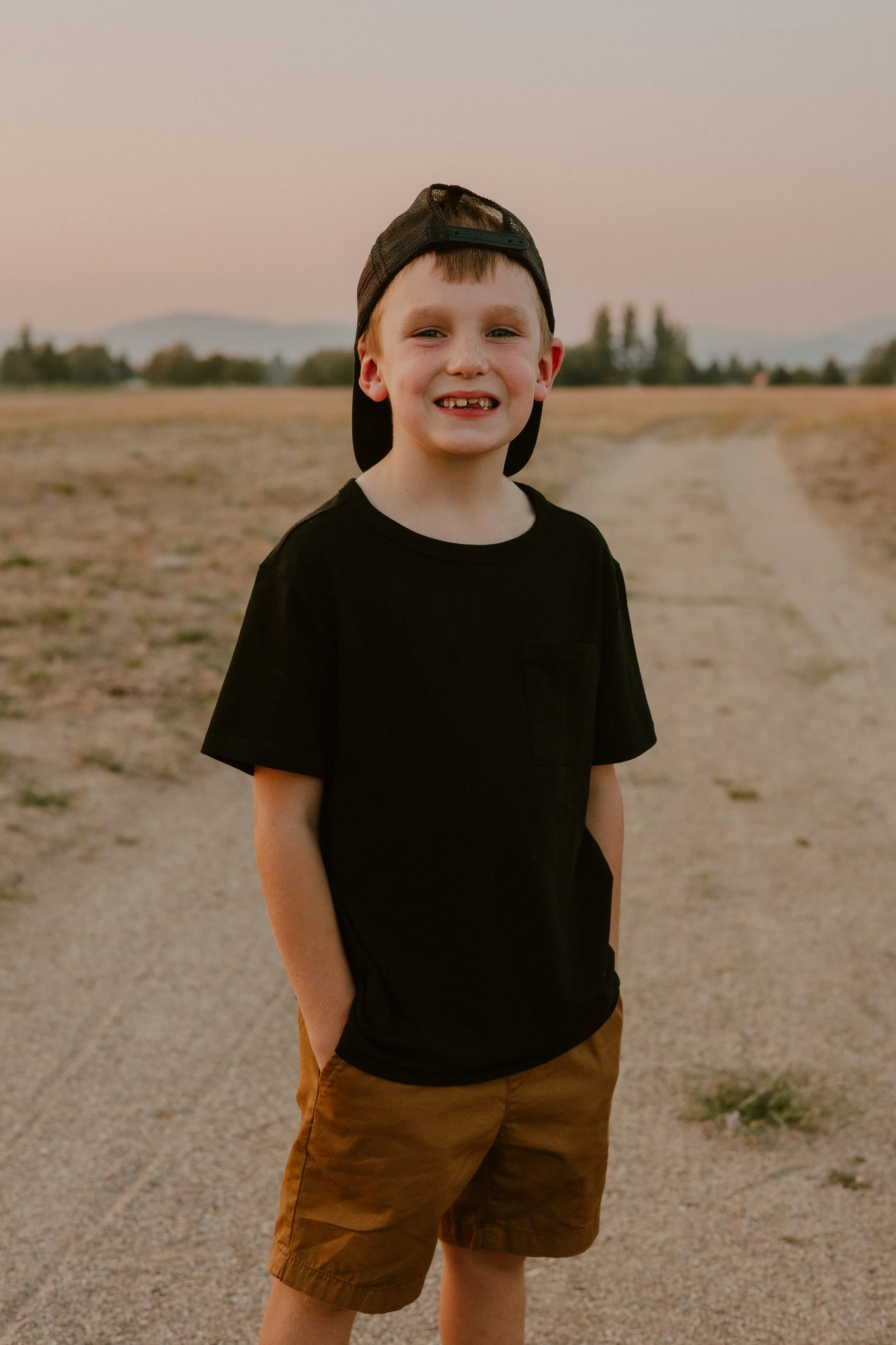 A young boy standing outdoors on a dirt pathway during sunset, wearing a black T-shirt, brown shorts, and a backward black cap, smiling at the camera.