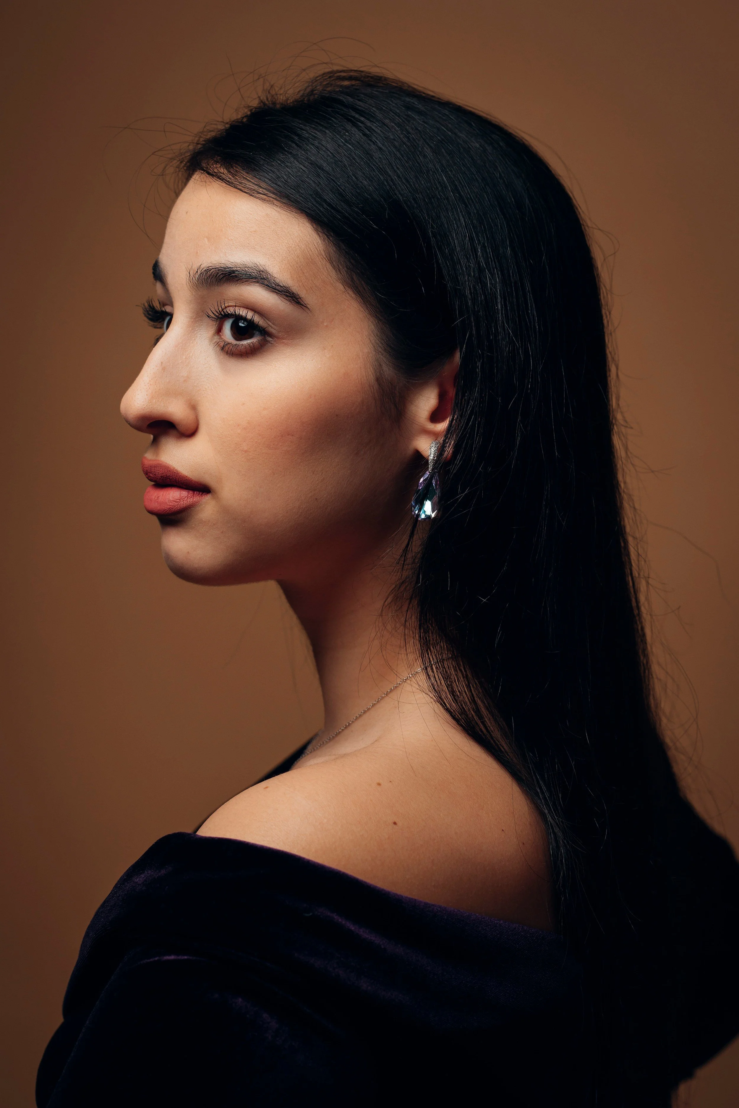 A woman with long black hair and earrings looking to her left against a brown background.