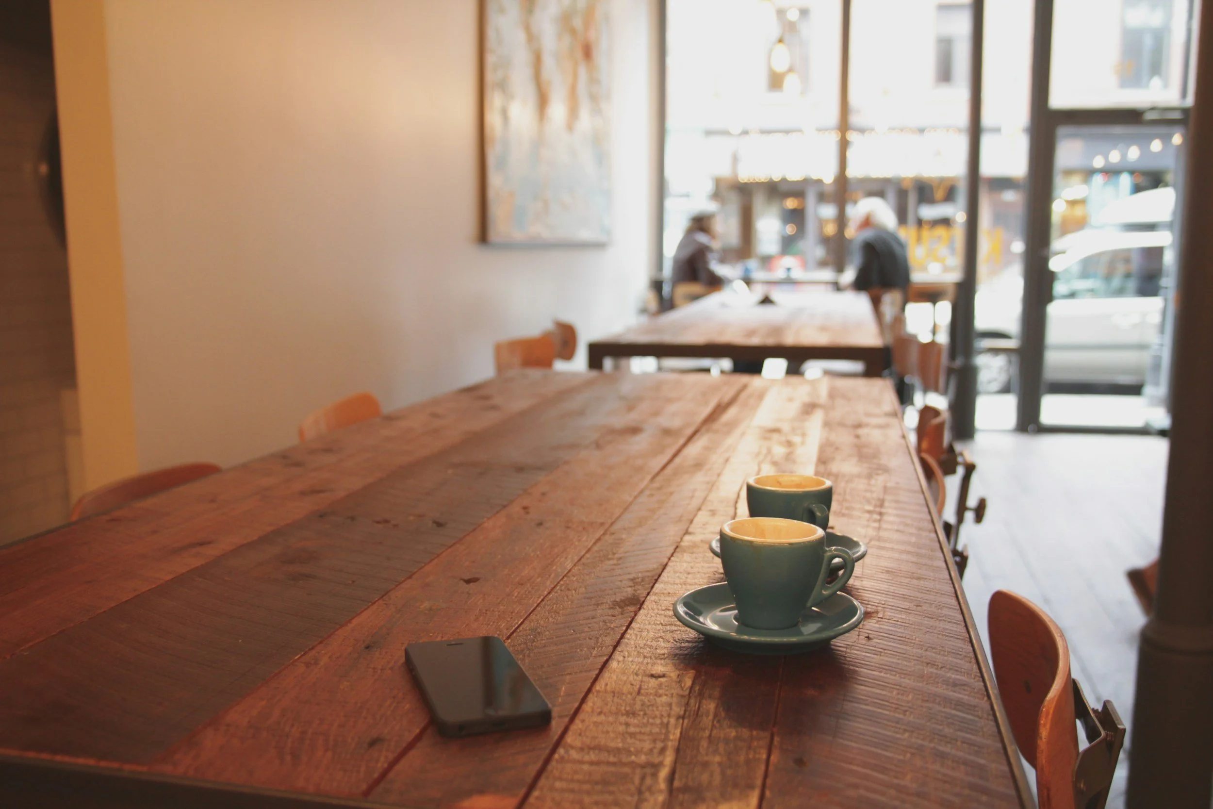 Interior of a cozy coffee shop with a long wooden table, two cups of coffee on saucers, and a smartphone, with large windows showing street view outside and a few customers inside.