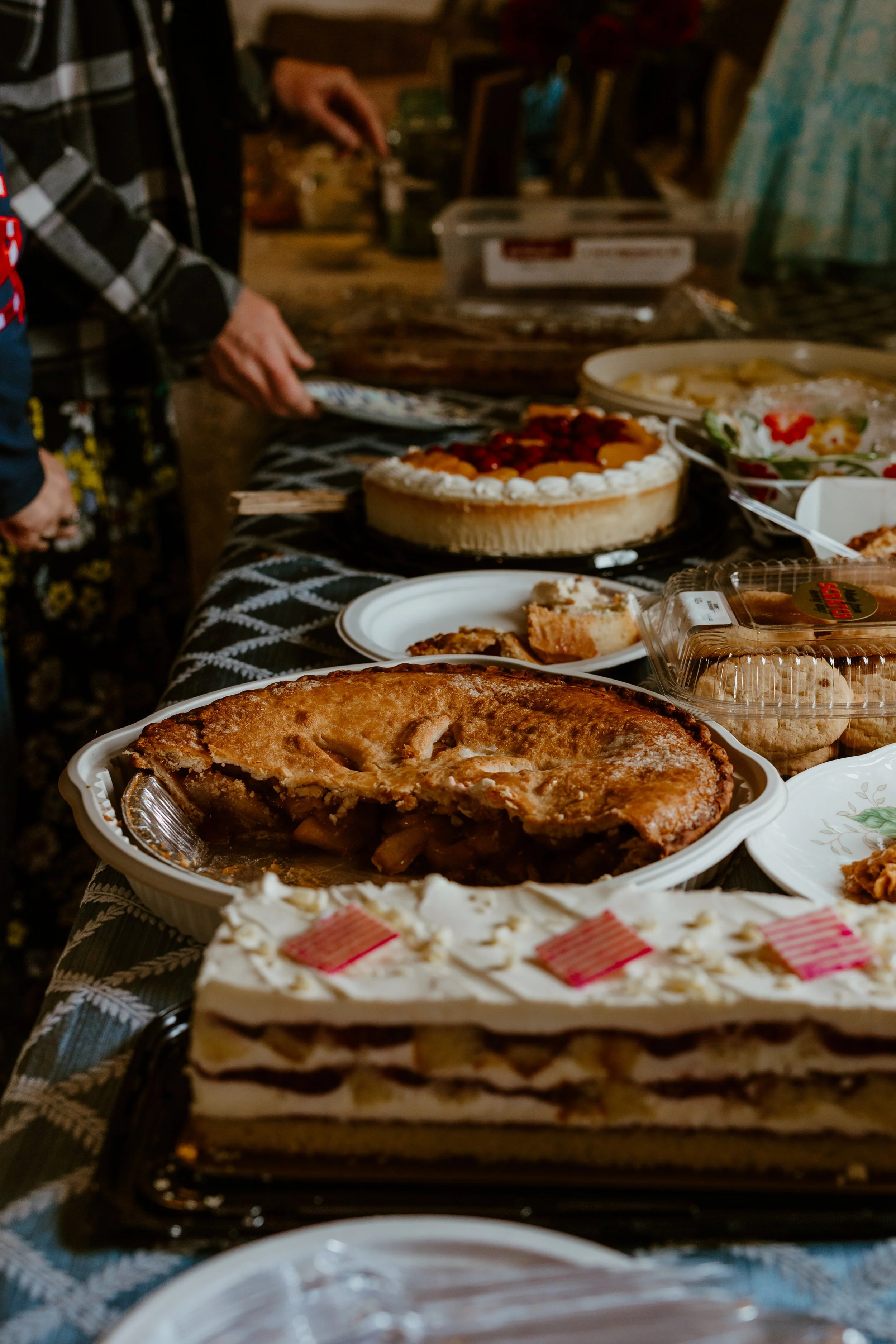 A variety of homemade pies and desserts on a table at a gathering. Visible are a cherry-topped pie, a lattice crust apple pie, a layer cake with white frosting and pink decorations, and other assorted sweets. People are seen in the background serving themselves.