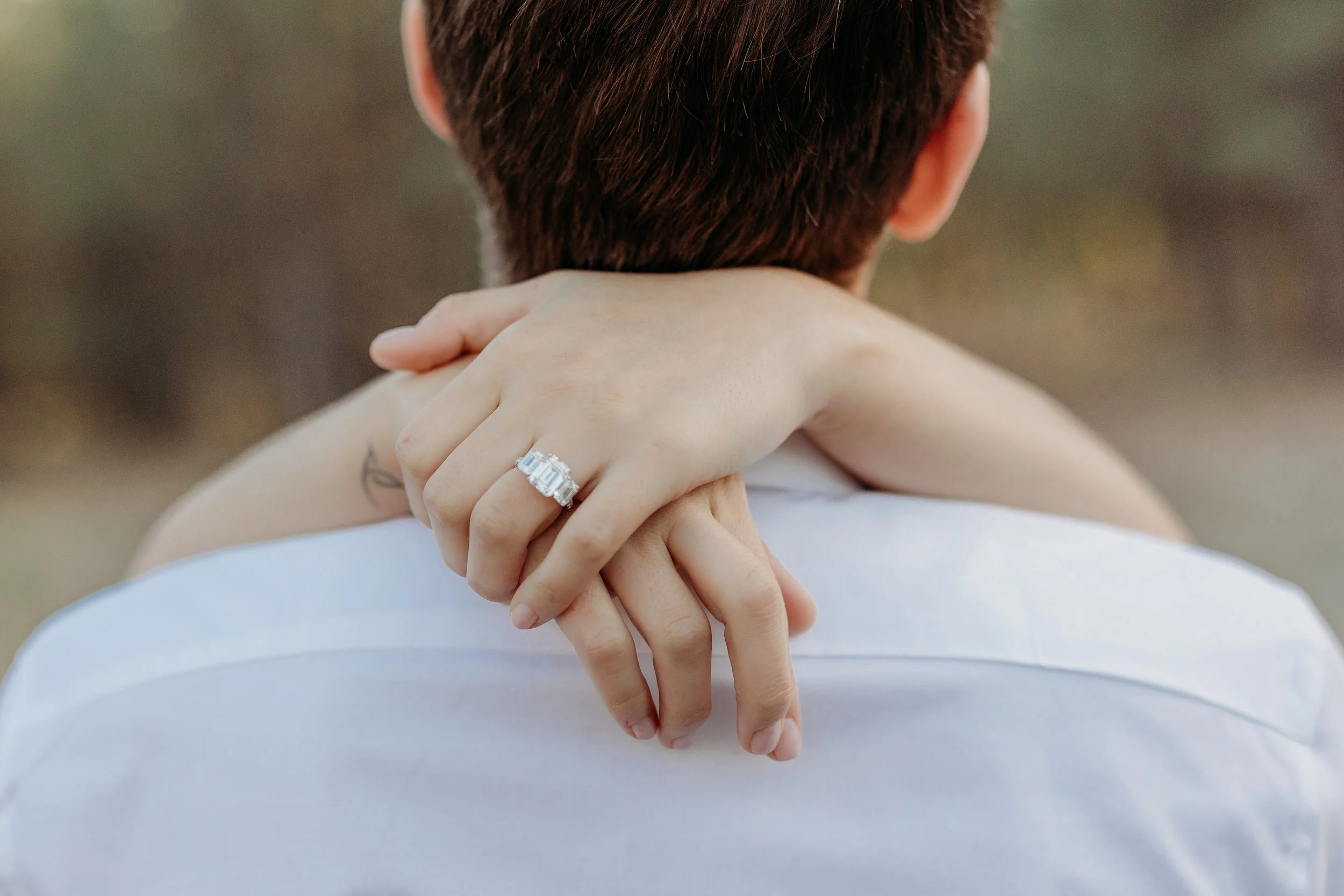 Close-up of a person with short brown hair, wearing a white shirt, holding their hands together behind their neck. One hand, with a large diamond engagement ring, rests on the other. The background is blurred with natural outdoor tones.