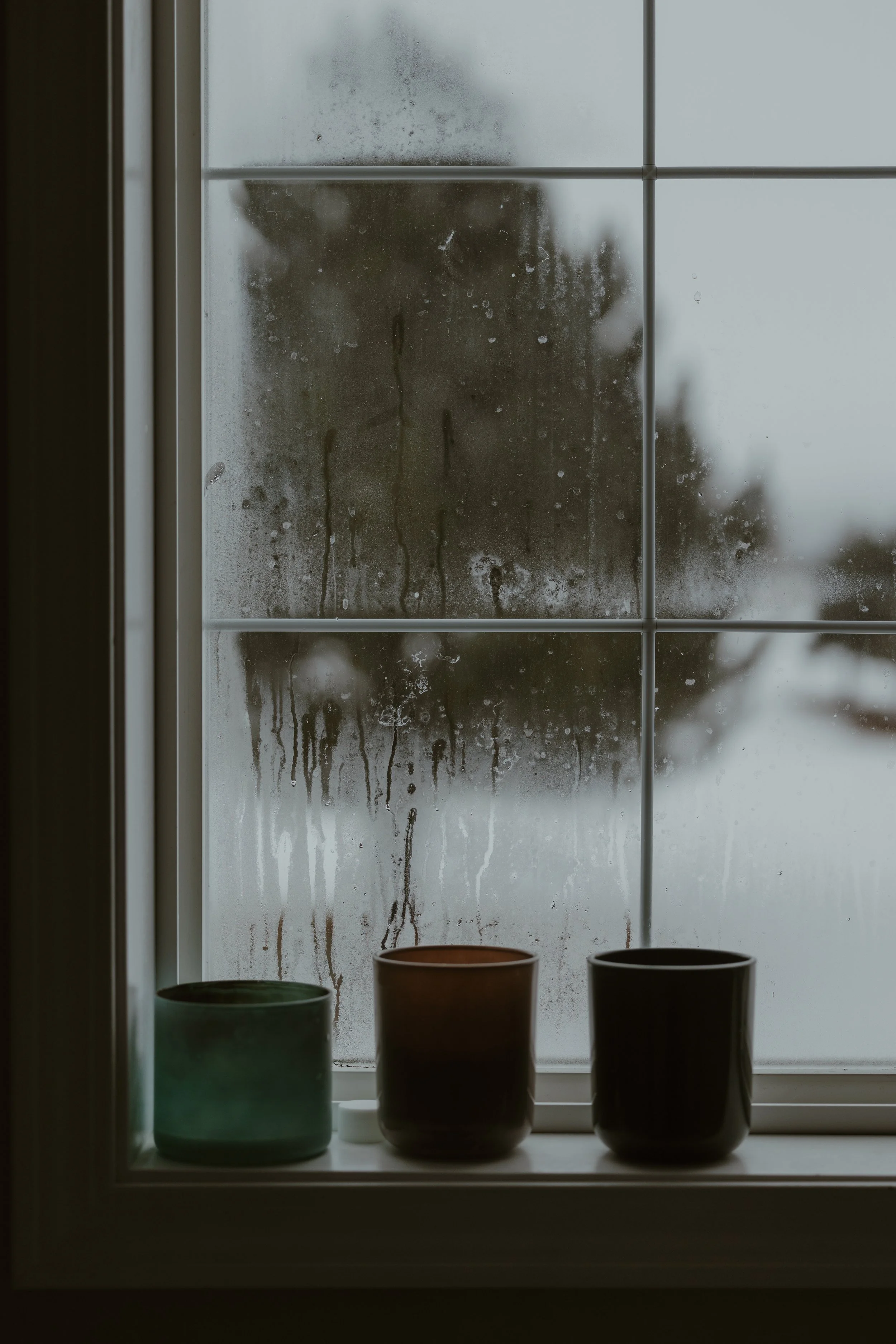 A window with condensation and water droplets, with three small cups on the windowsill in front of it.