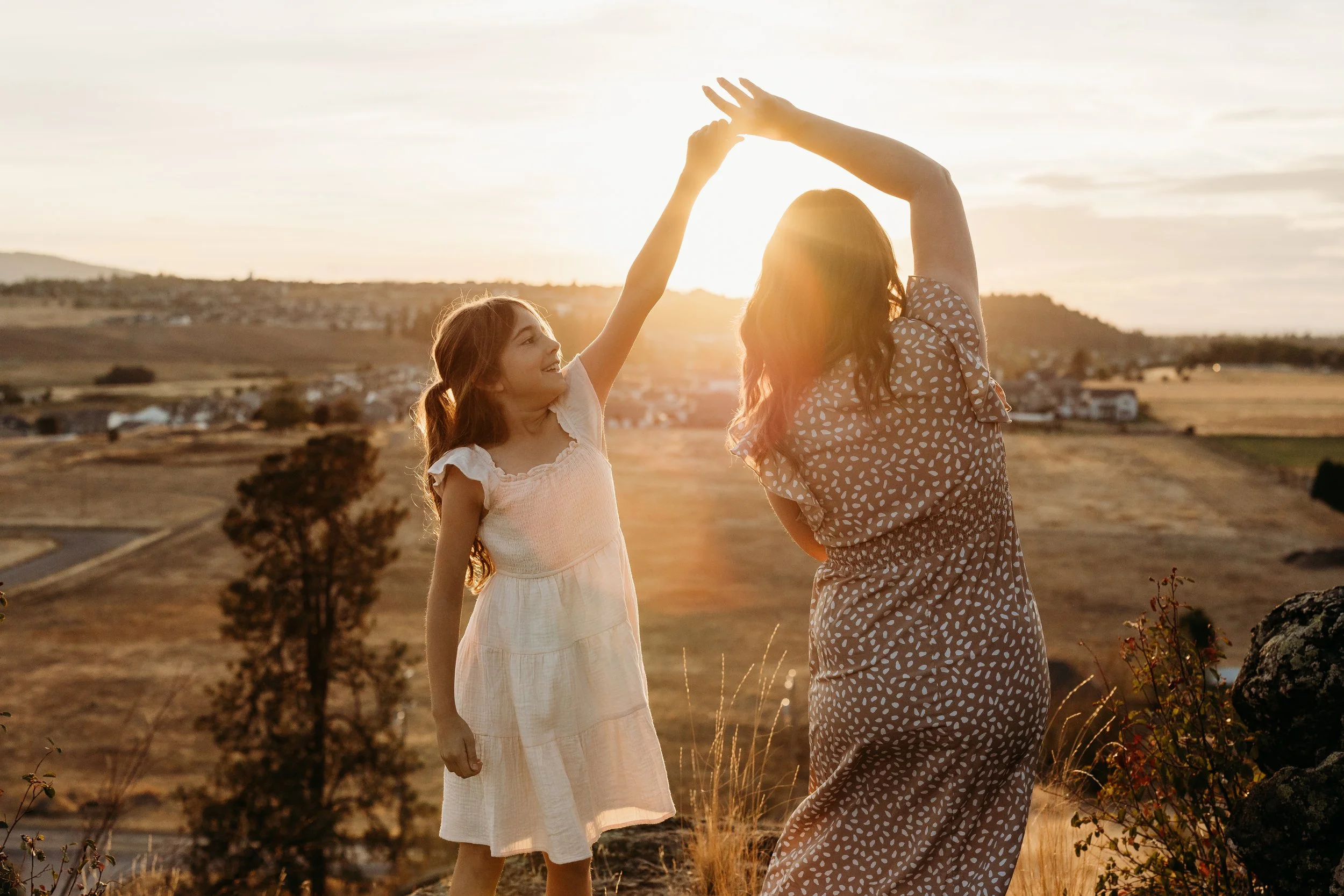 A woman and a girl dancing outside during sunset, with open fields and a small town in the background.