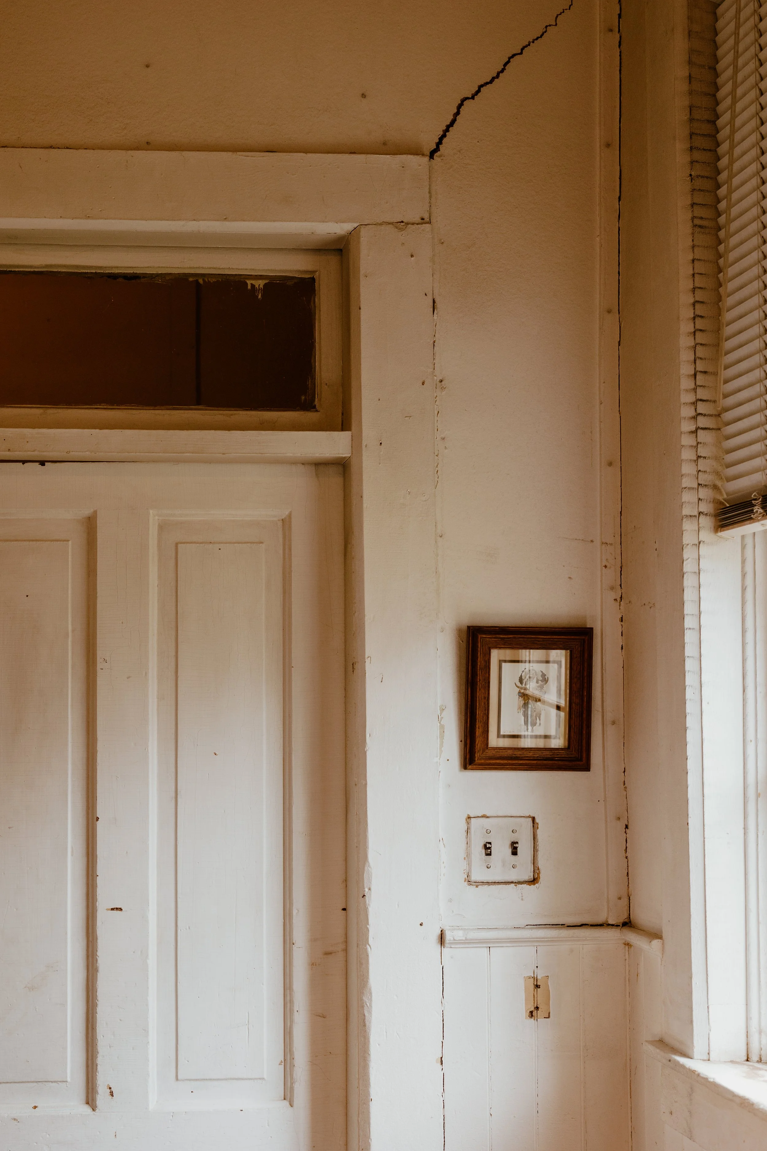 Interior wall with damaged paint and wiring switch, framed picture, and window with blinds.