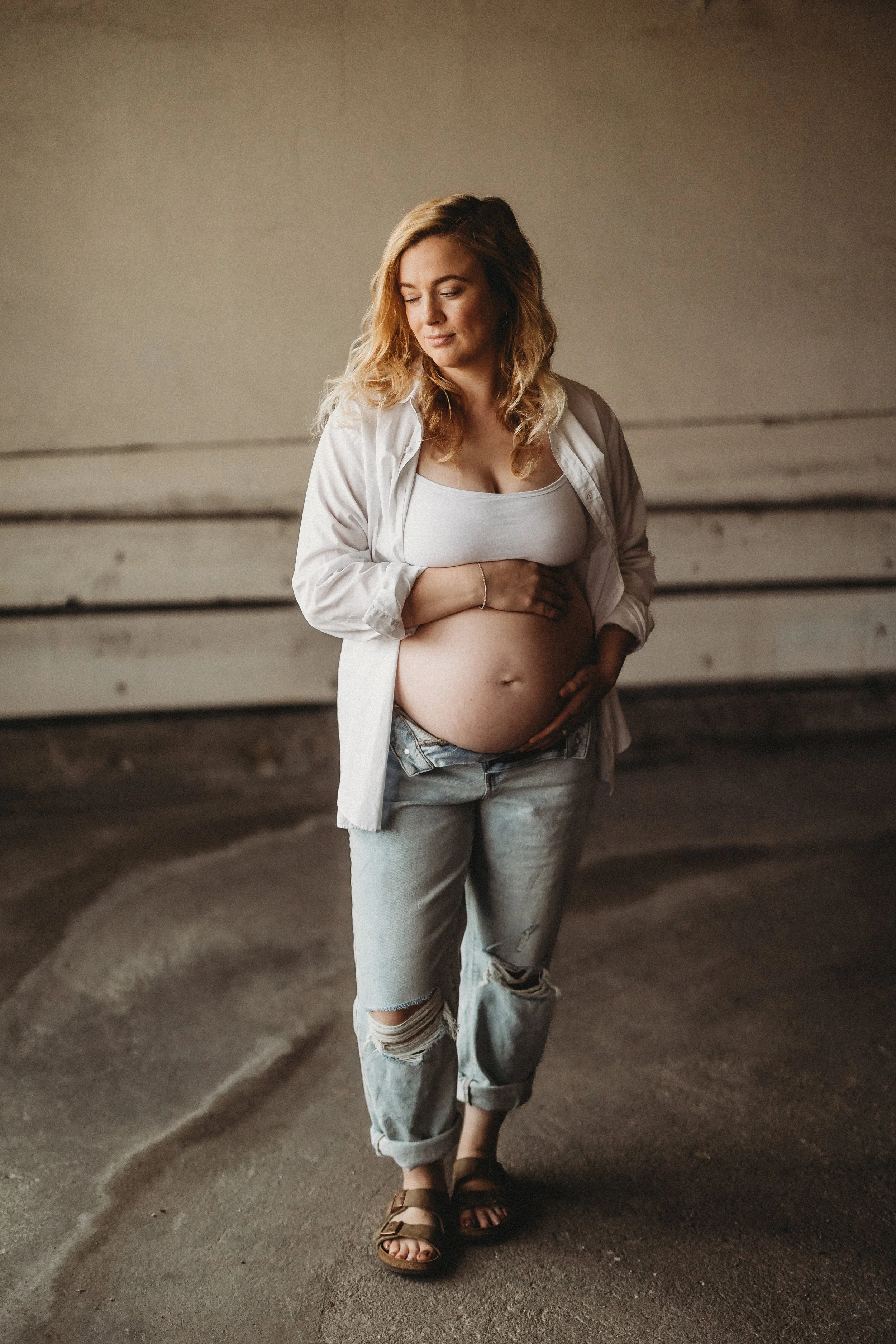 Pregnant woman standing outdoors, gently holding her baby bump and looking down, wearing casual clothing.