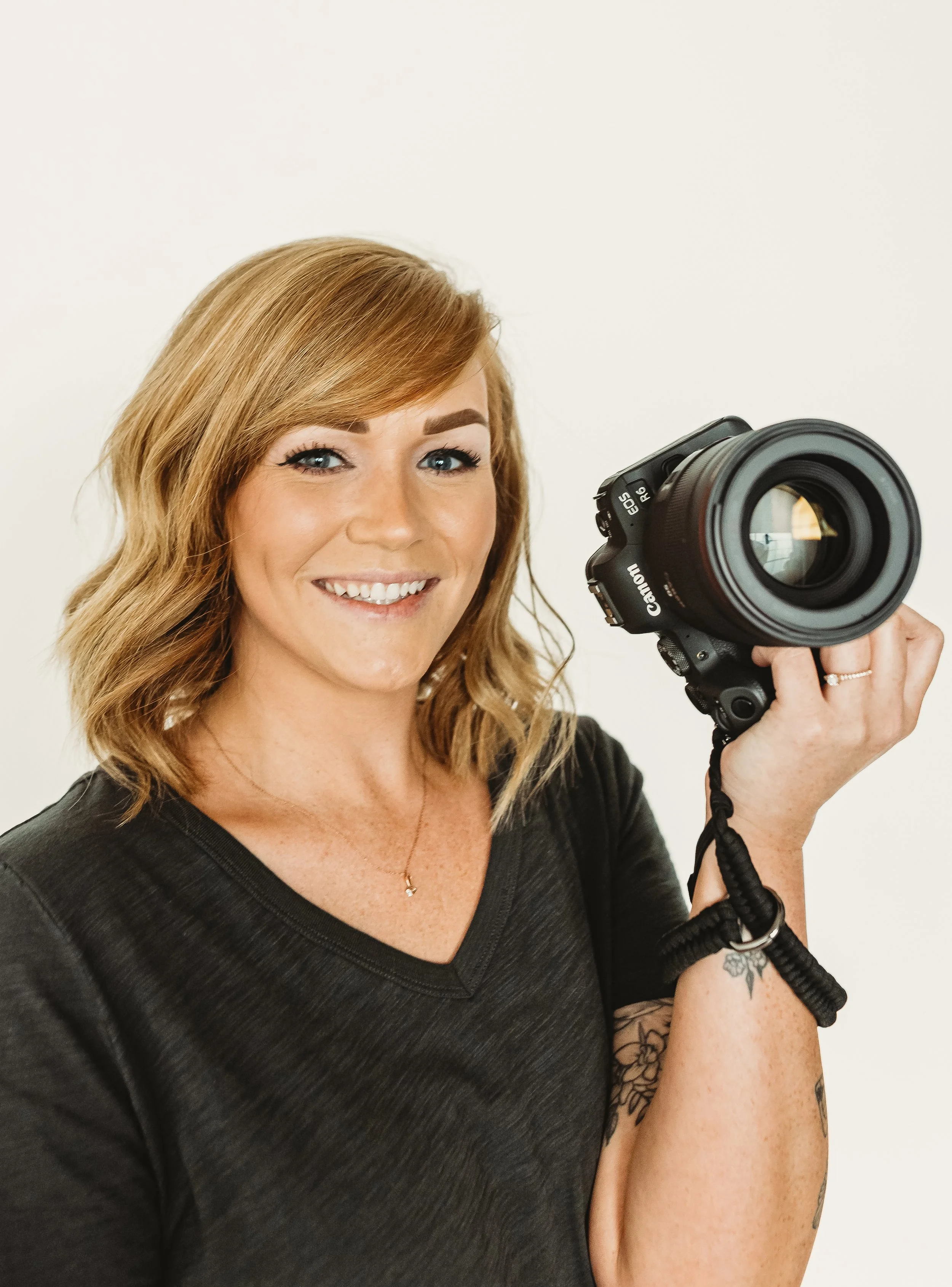 A woman with light brown wavy hair holding a camera, smiling, wearing a black V-neck shirt and a gold necklace, against a plain white background.