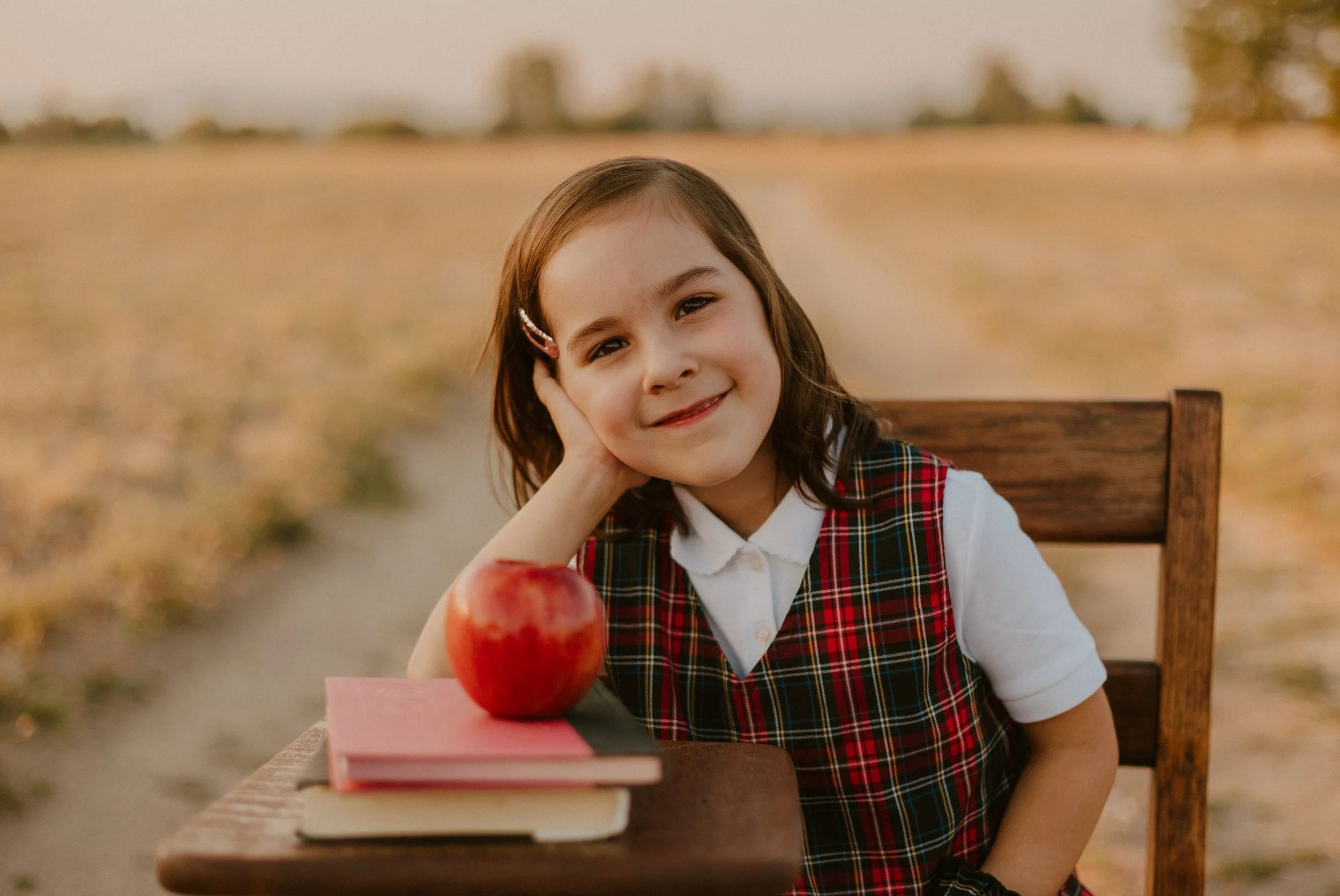 A young girl with brown hair wearing a plaid dress sits at a wooden table outdoors. She rests her head on her hand and smiles. In front of her are two books and a red apple.