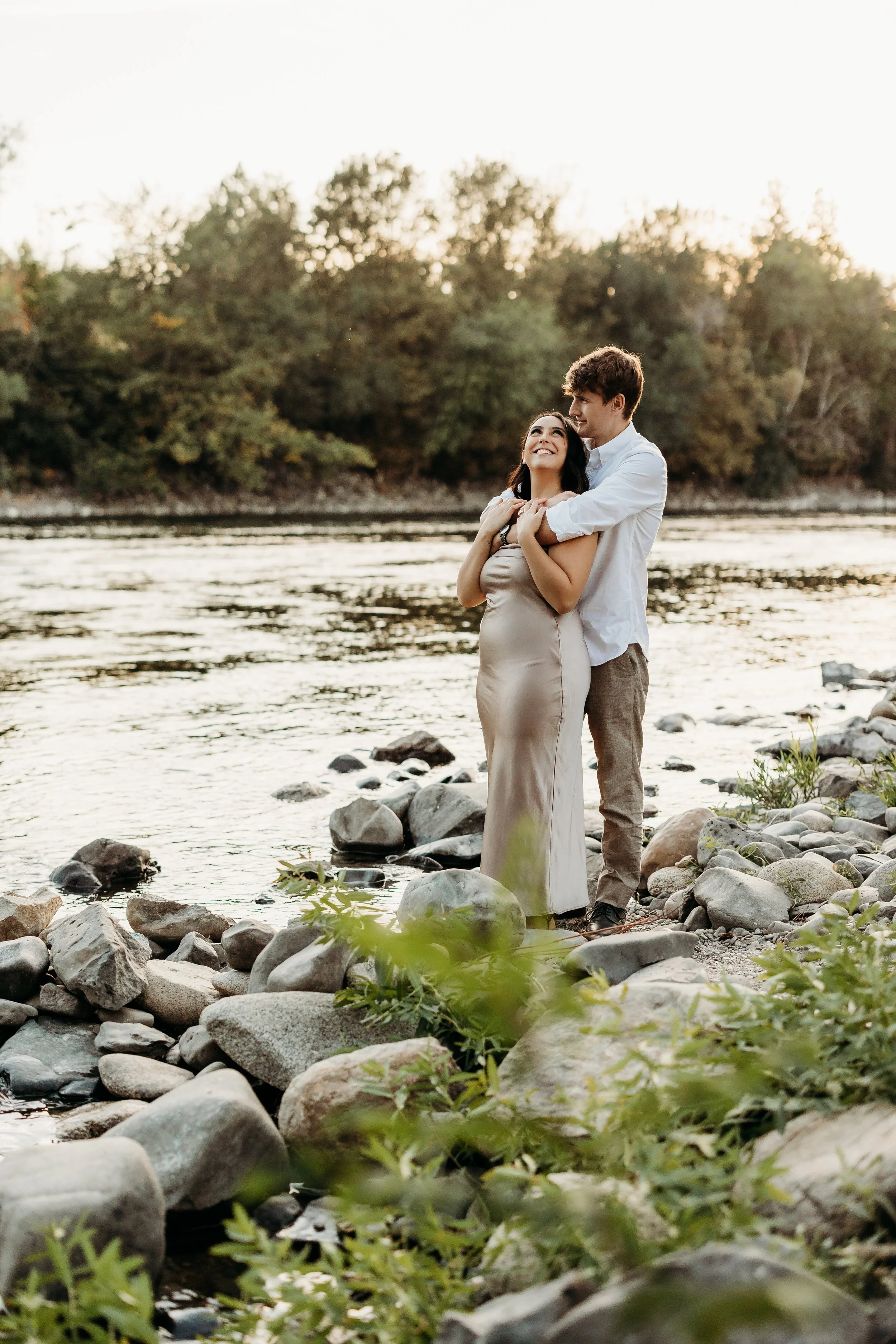 A couple standing by a river with trees in the background, embracing and smiling at each other during sunset.