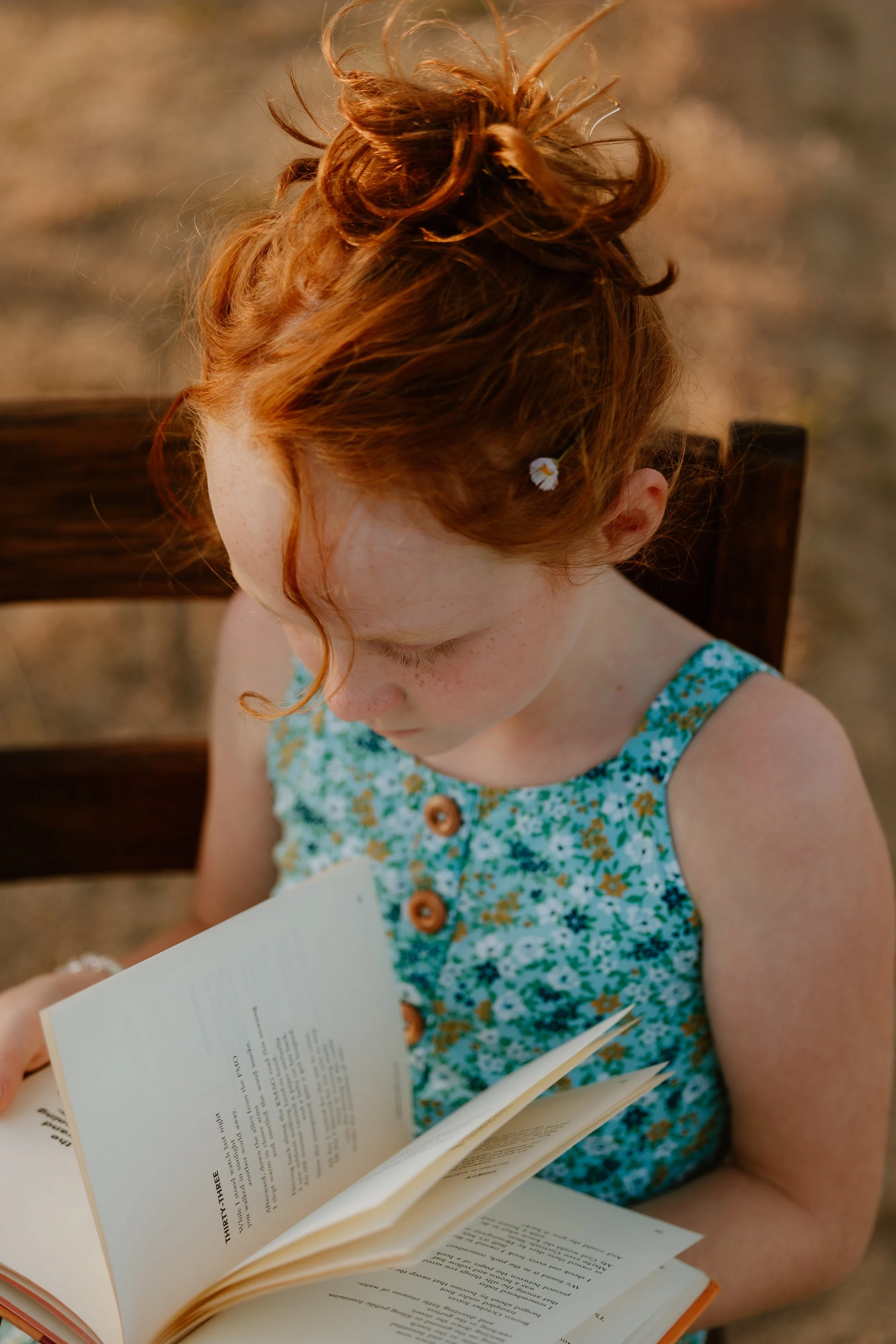 A young girl with red hair and freckles reads a book while sitting outdoors on a wooden chair, wearing a blue floral dress.