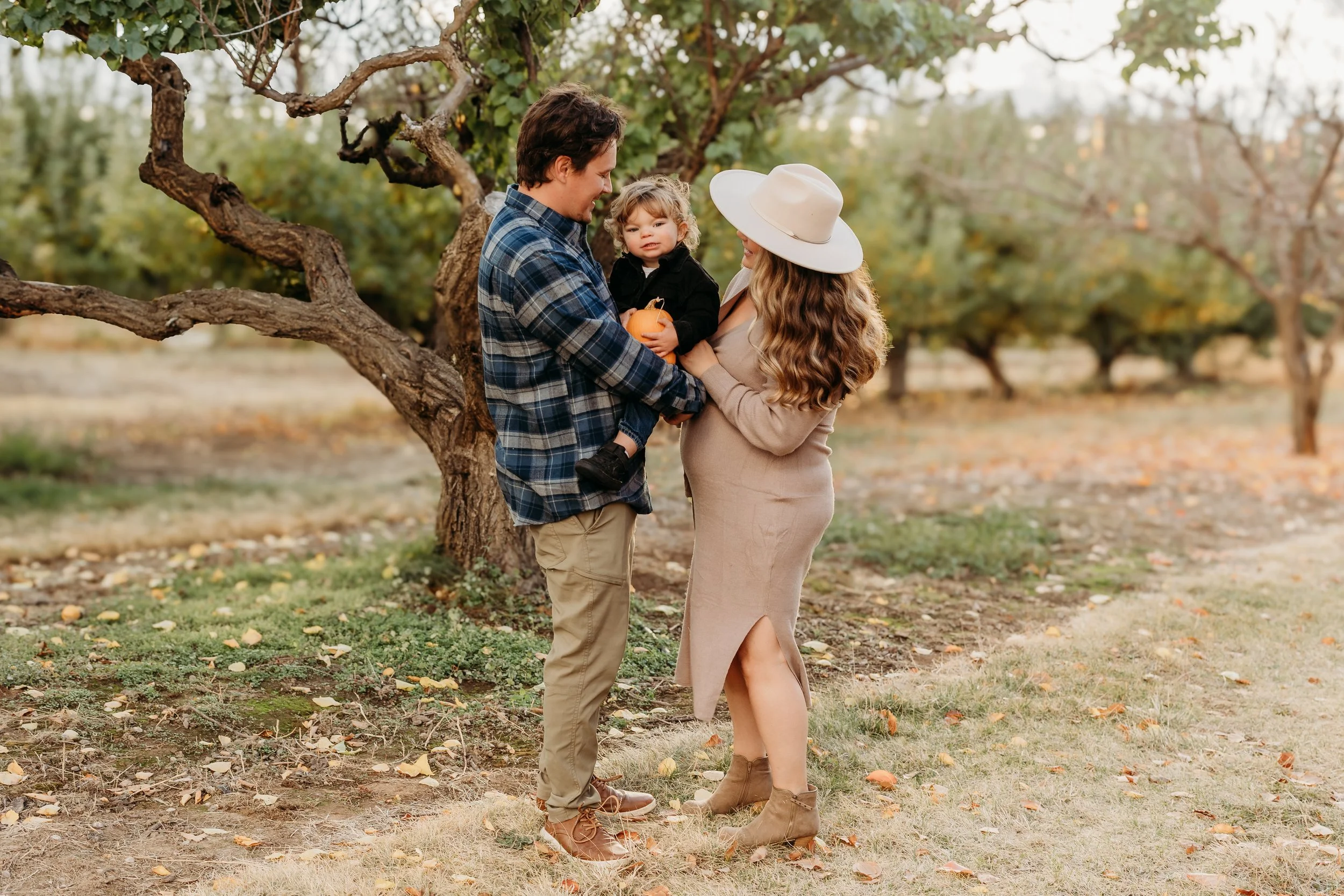 A family of three outdoors near a tree, with autumn leaves on the ground: a man, woman, and young girl holding a small pumpkin, smiling and interacting.
