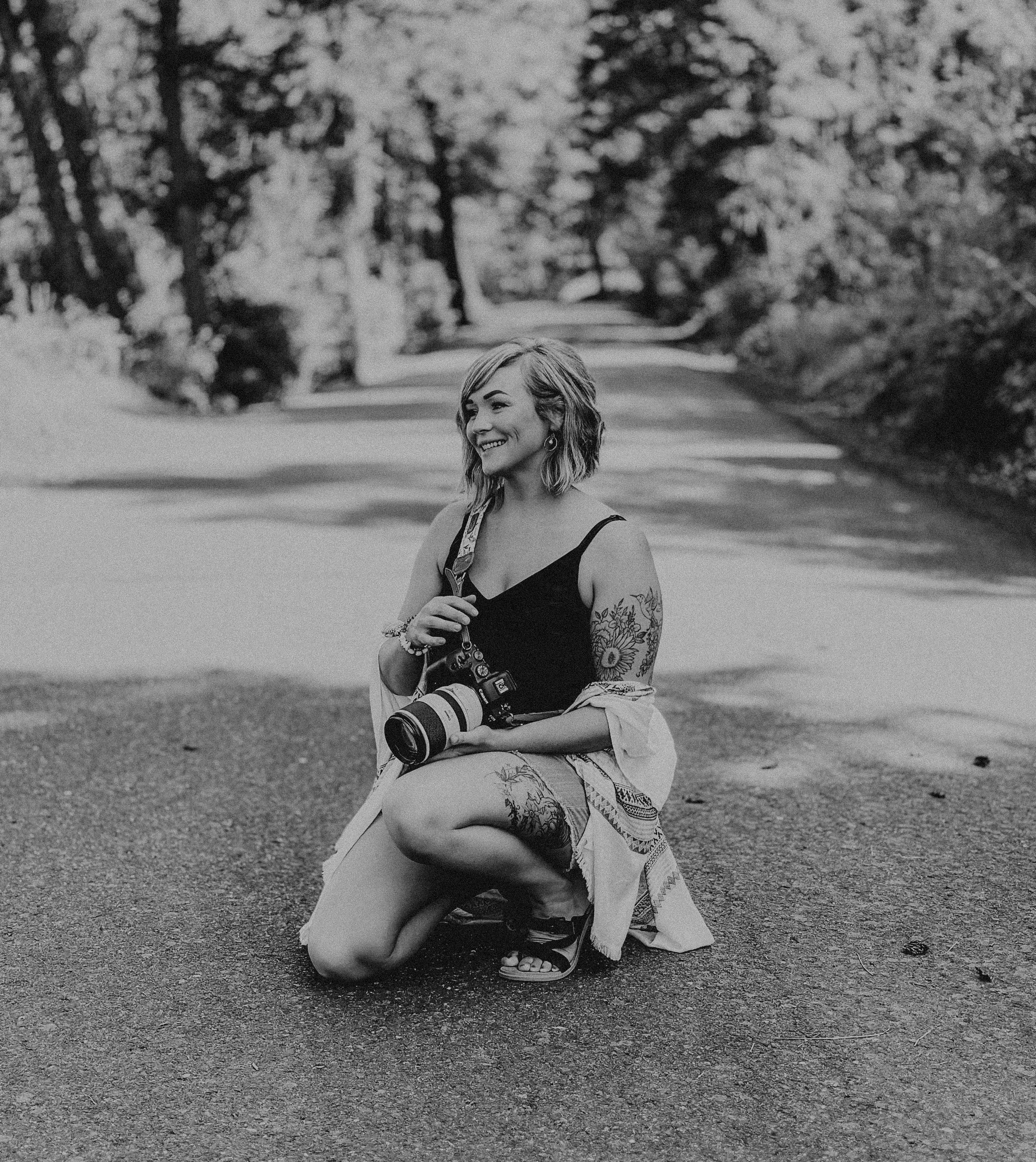 A woman with tattoos on her arm and leg, holding a camera, kneels on a dirt path in a wooded area, smiling and looking to her left.