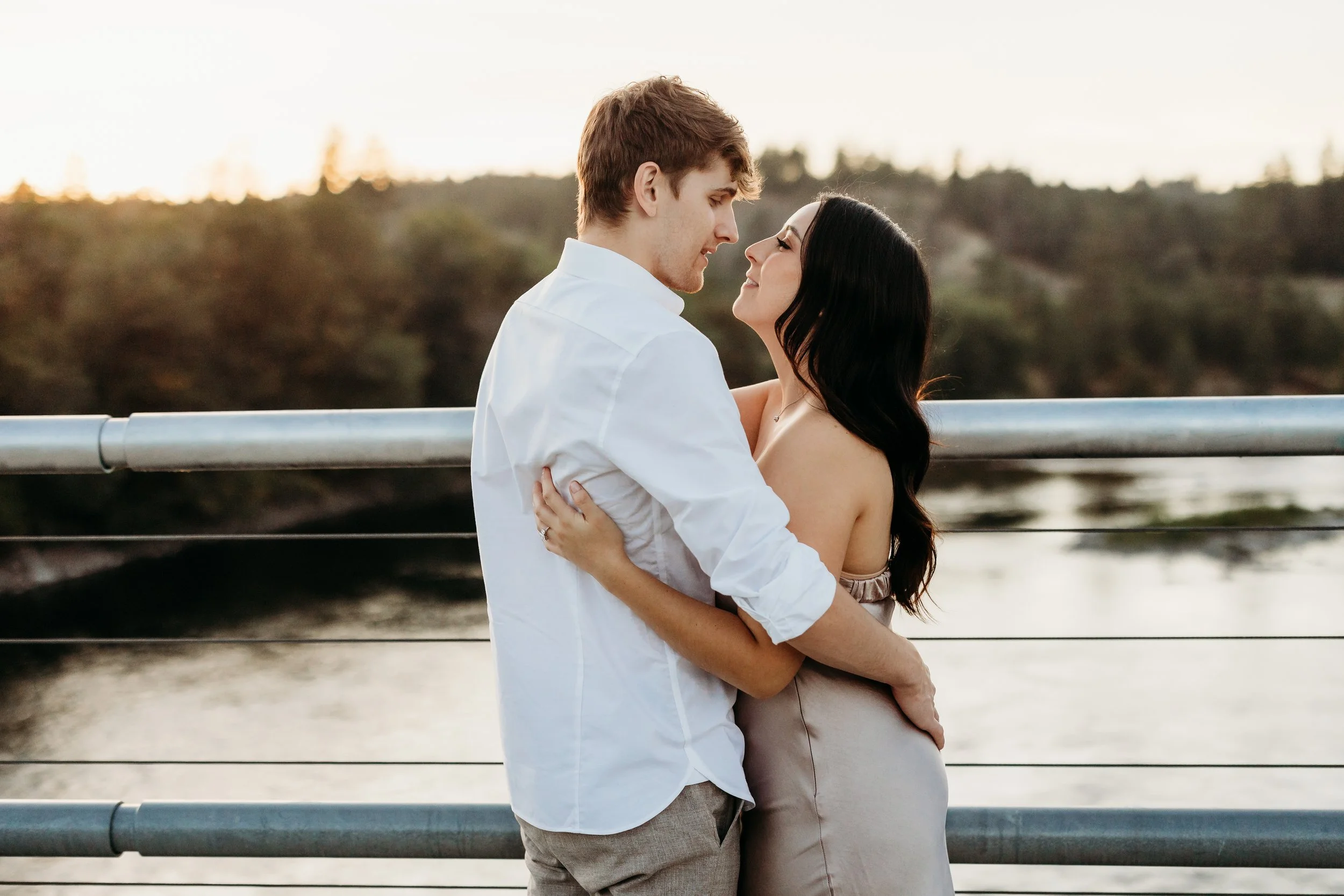 A couple with light skin and dark hair embracing on a bridge over water during sunset.
