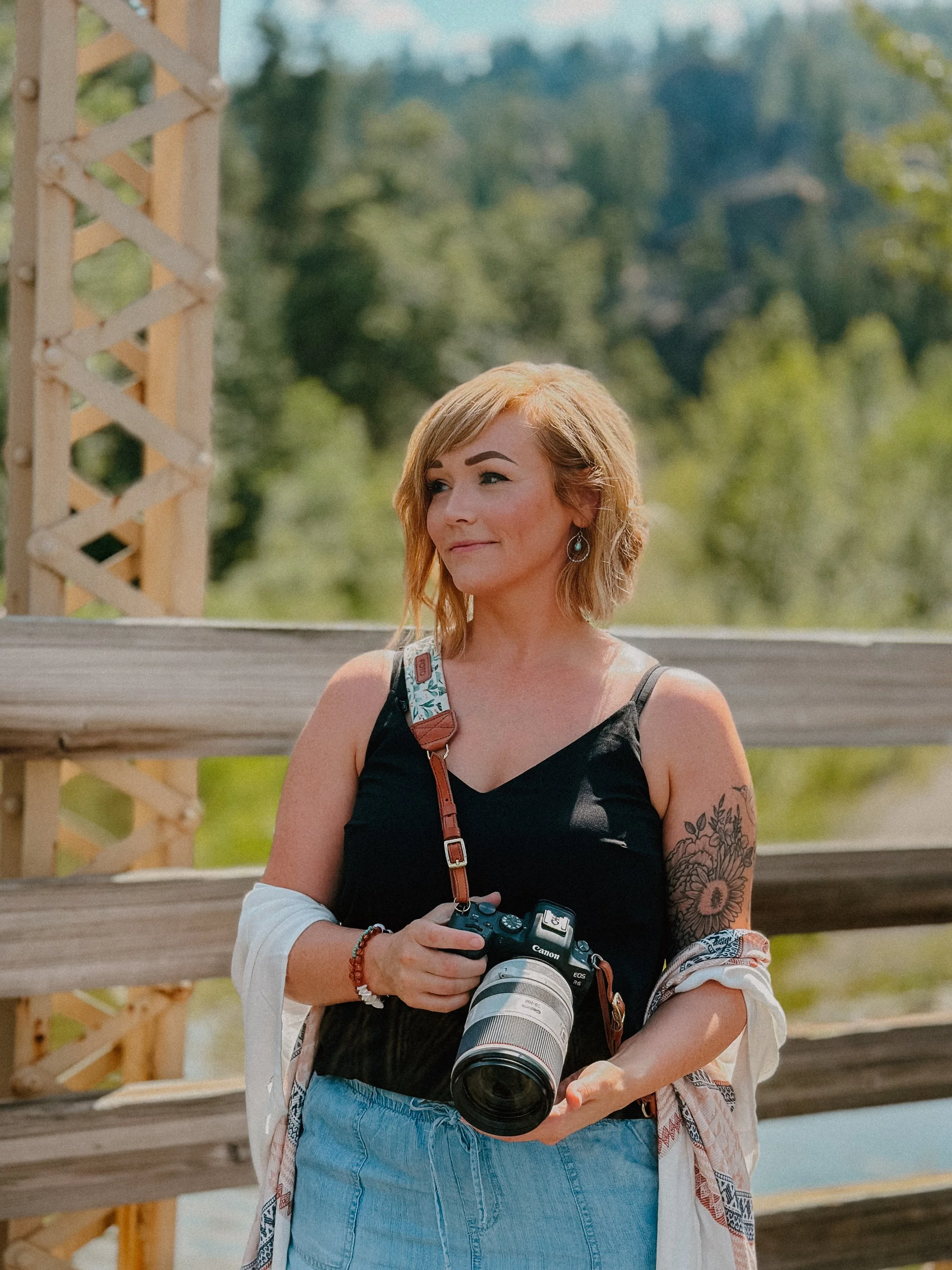 A woman with short, wavy blonde hair holding a camera, standing outdoors with a wooden railing and lush green trees in the background.