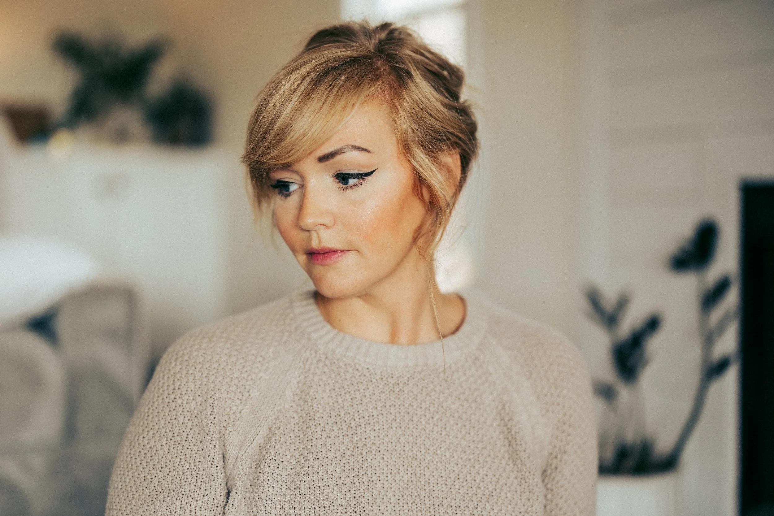 A woman with short blonde hair and winged eyeliner looking down, wearing a beige knit sweater, in a cozy indoor setting with blurred background decor.