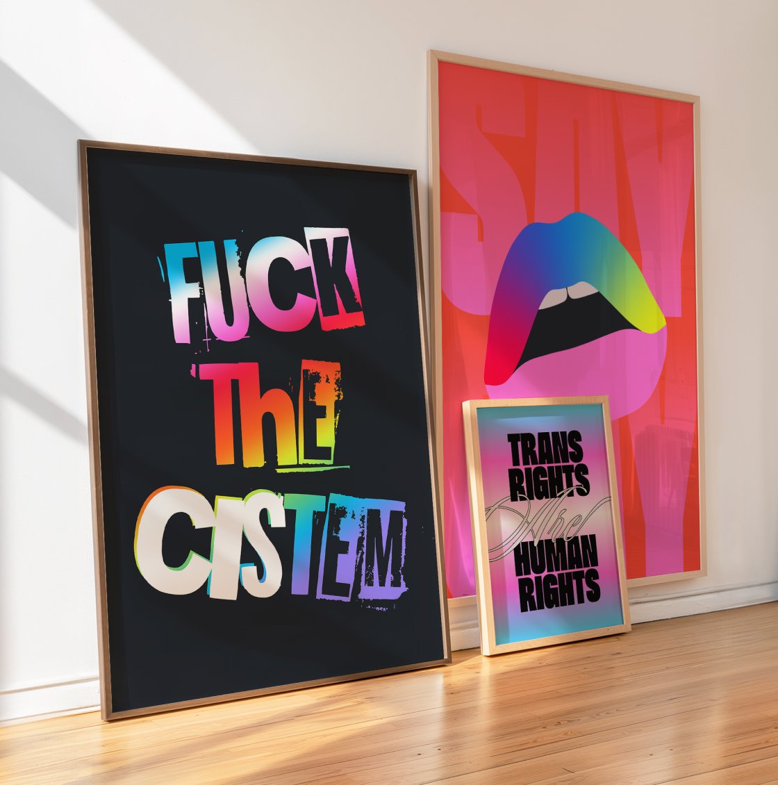 Three colorful posters leaning against a wall. The first poster reads "FUCK THE SYSTEM" in rainbow-colored graffiti-style text on a black background. The second poster features a stylized illustration of lips with a rainbow gradient. The third poster says "TRANS RIGHTS ARE HUMAN RIGHTS" in bold black letters on a pastel gradient background.