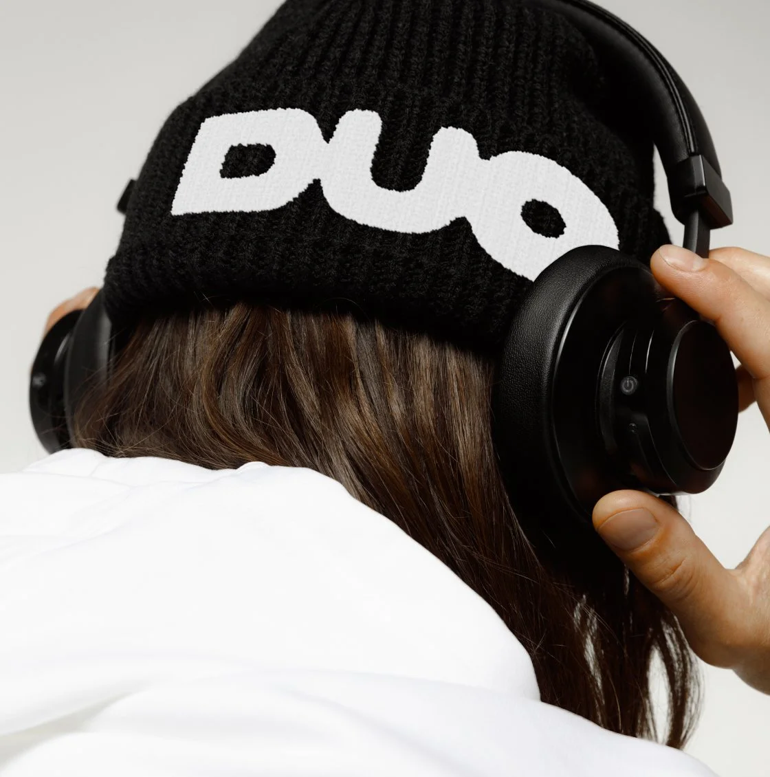 A close-up of a person wearing a black knit beanie with the DUO logo, paired with sleek headphones, capturing a modern urban style and branding integration.