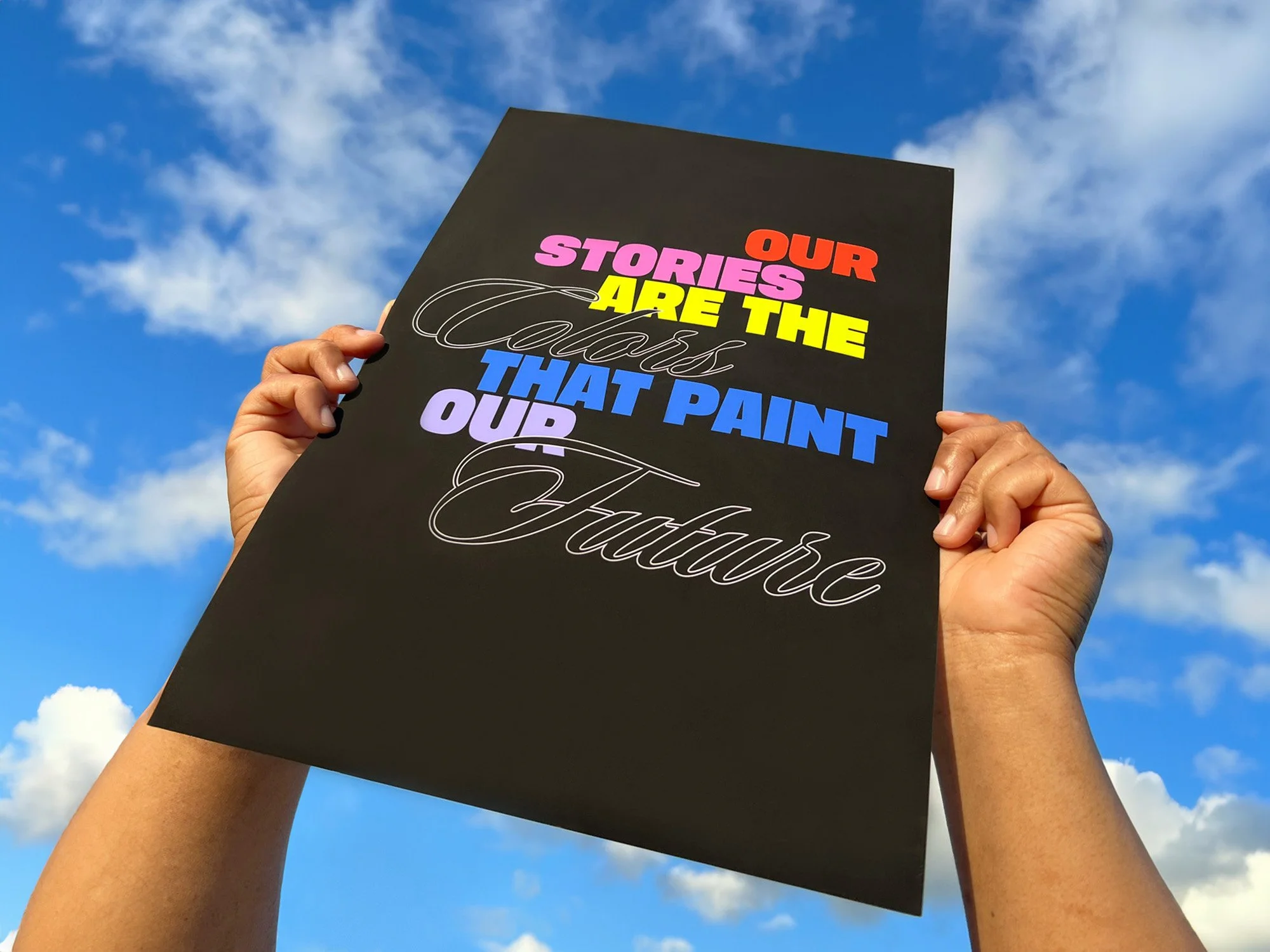 Person holding a black sign with colorful text in front of a blue sky with clouds. The sign reads 'Our stories are the class that paint our futures'.