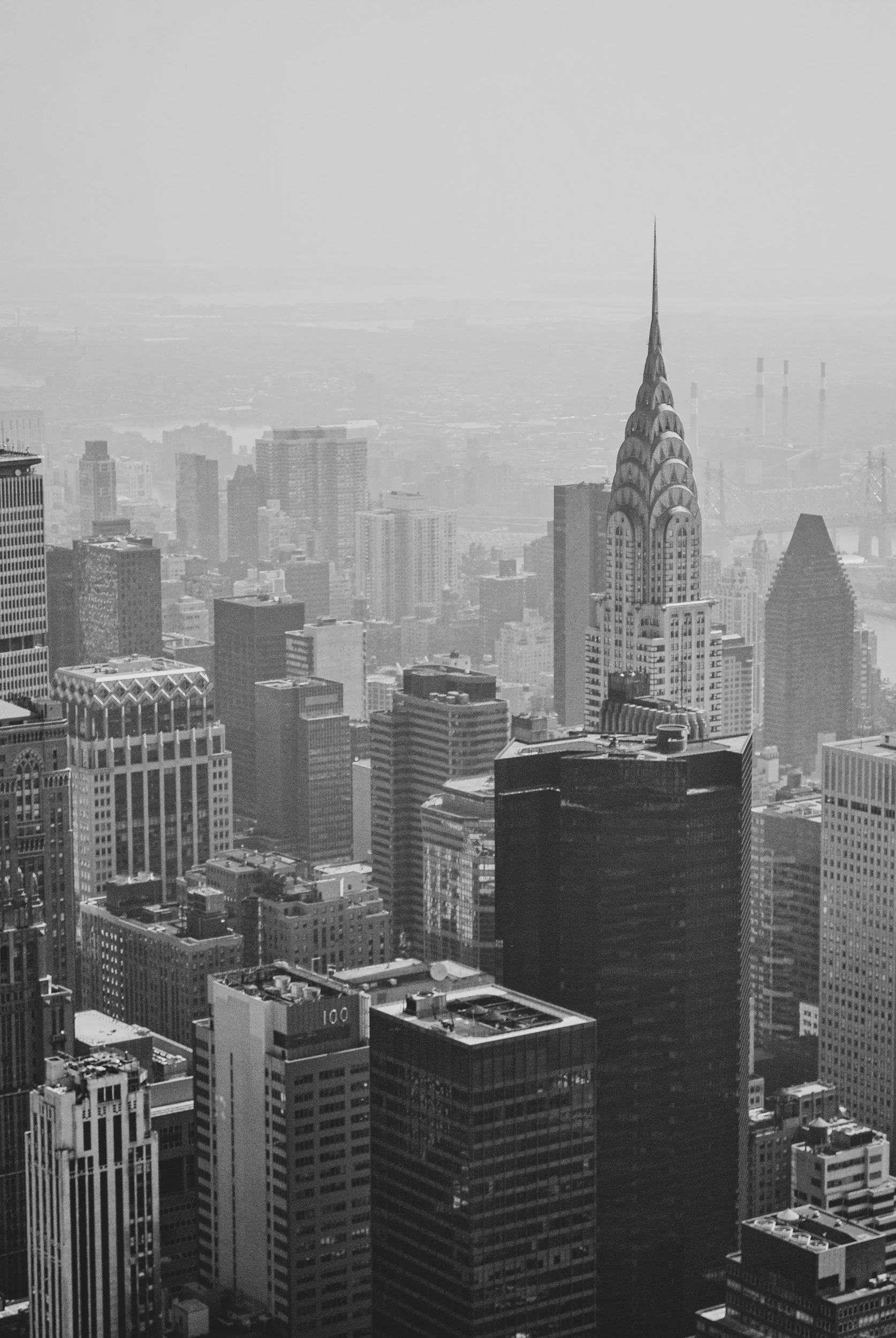 Black and white aerial view of a city skyline featuring the Chrysler Building with its distinctive art deco spire, surrounded by numerous tall skyscrapers, with a hazy background and industrial chimneys in the distance.