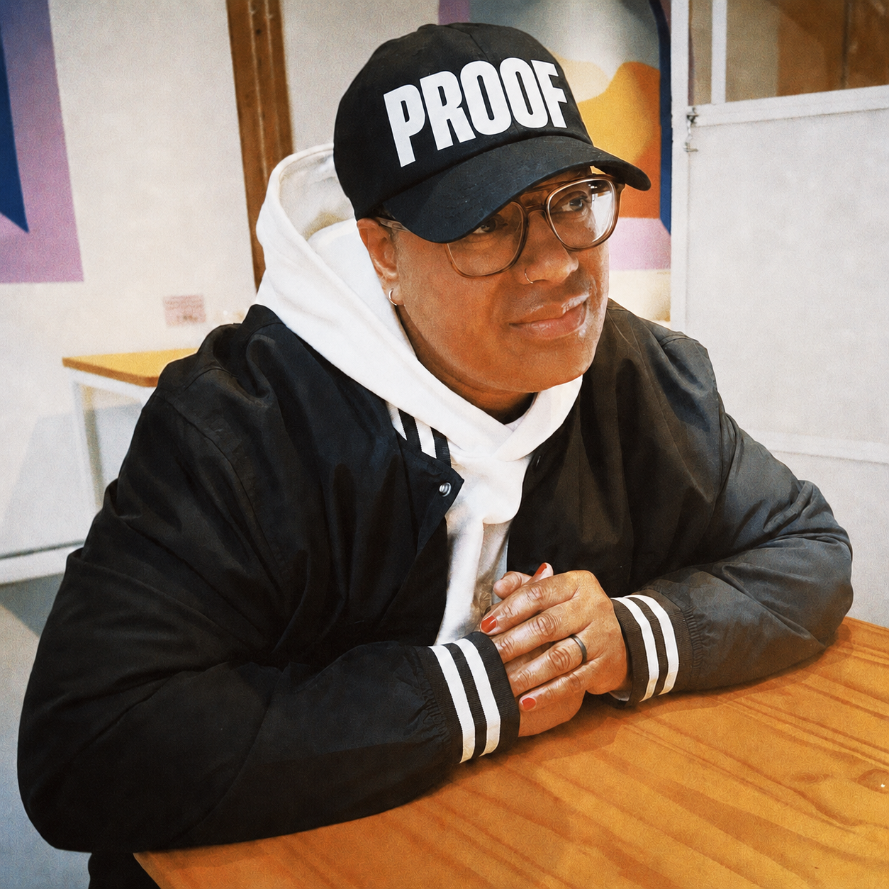 A person sitting at a wooden table wearing glasses, a black cap with the word 'PROOF' on it, a black jacket, and a white hoodie. Hands are clasped on the table.