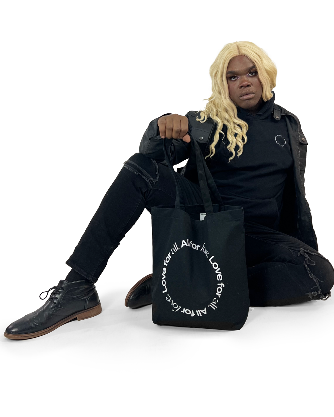 Person with long blonde hair, black clothing, and black shoes sitting on the floor, holding a black bag with white circular text that reads "Love for all. All for love."