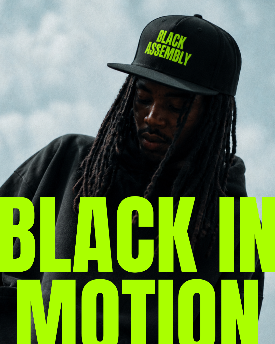 A person with long dreadlocks wearing a black cap with bright green text that reads "Black Assembly" and a black jacket, looking down against a cloudy sky background. Large bright green text says "Black in Motion" across the image.