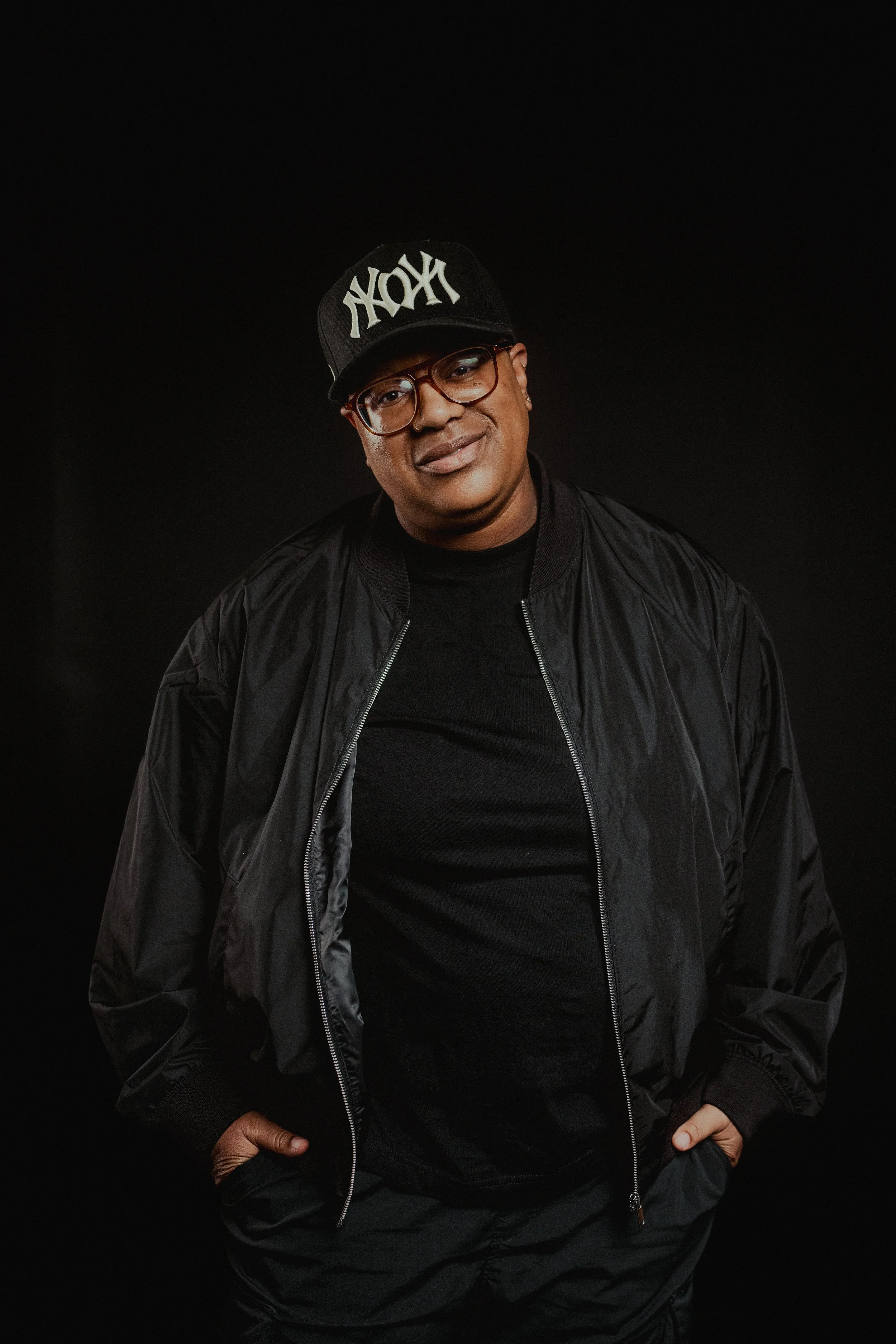 A man wearing glasses, a black jacket, a black cap with white lettering, and black pants, standing against a black background.
