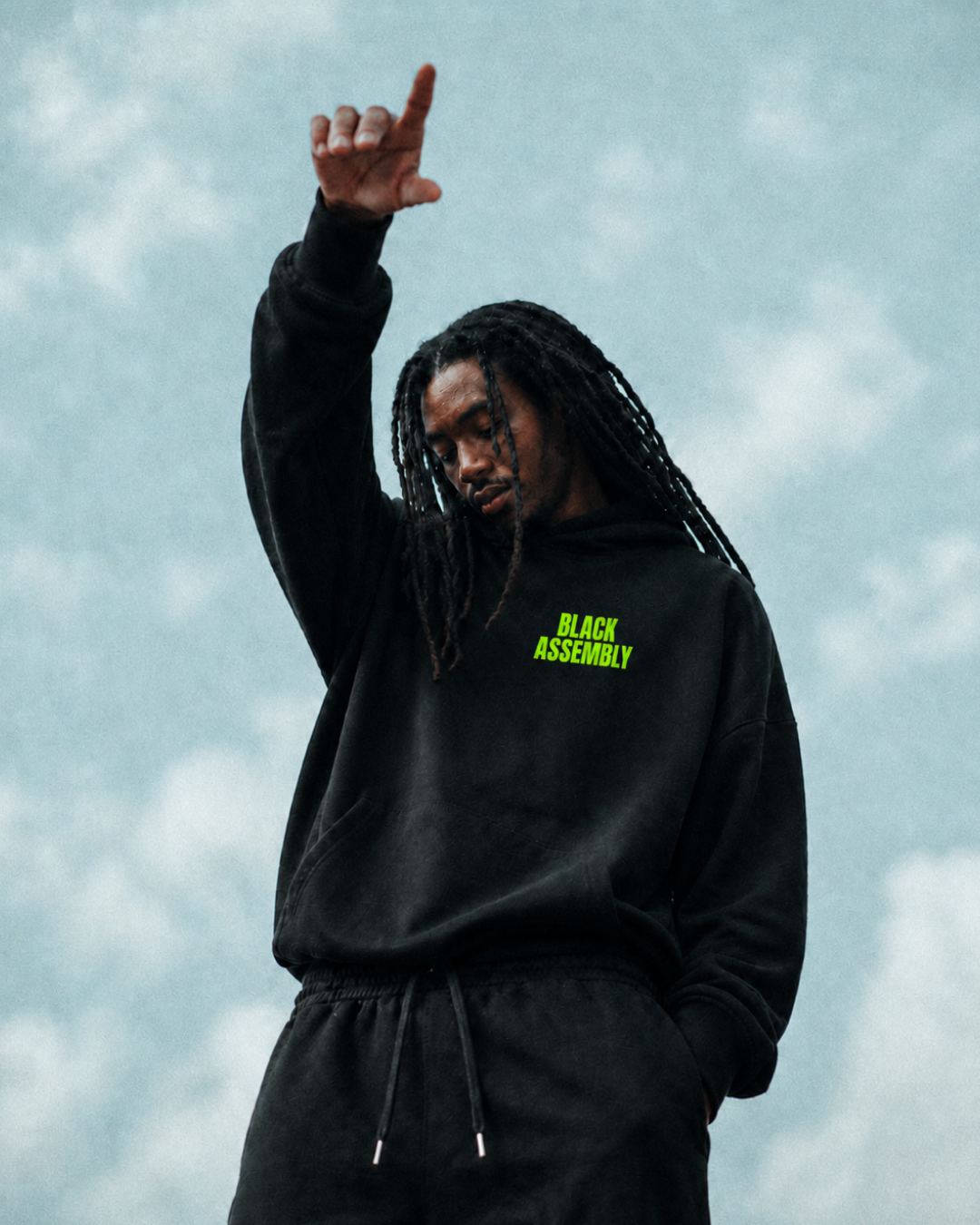 A young man with dreadlocks wearing a black hoodie with yellow-green 'Black Assembly' text, standing outdoors under cloudy sky, looking down with one hand raised.
