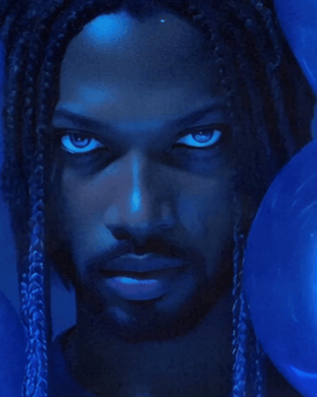 Close-up of a person's face with dark skin, intense expression, and braided hair, illuminated by blue lighting.
