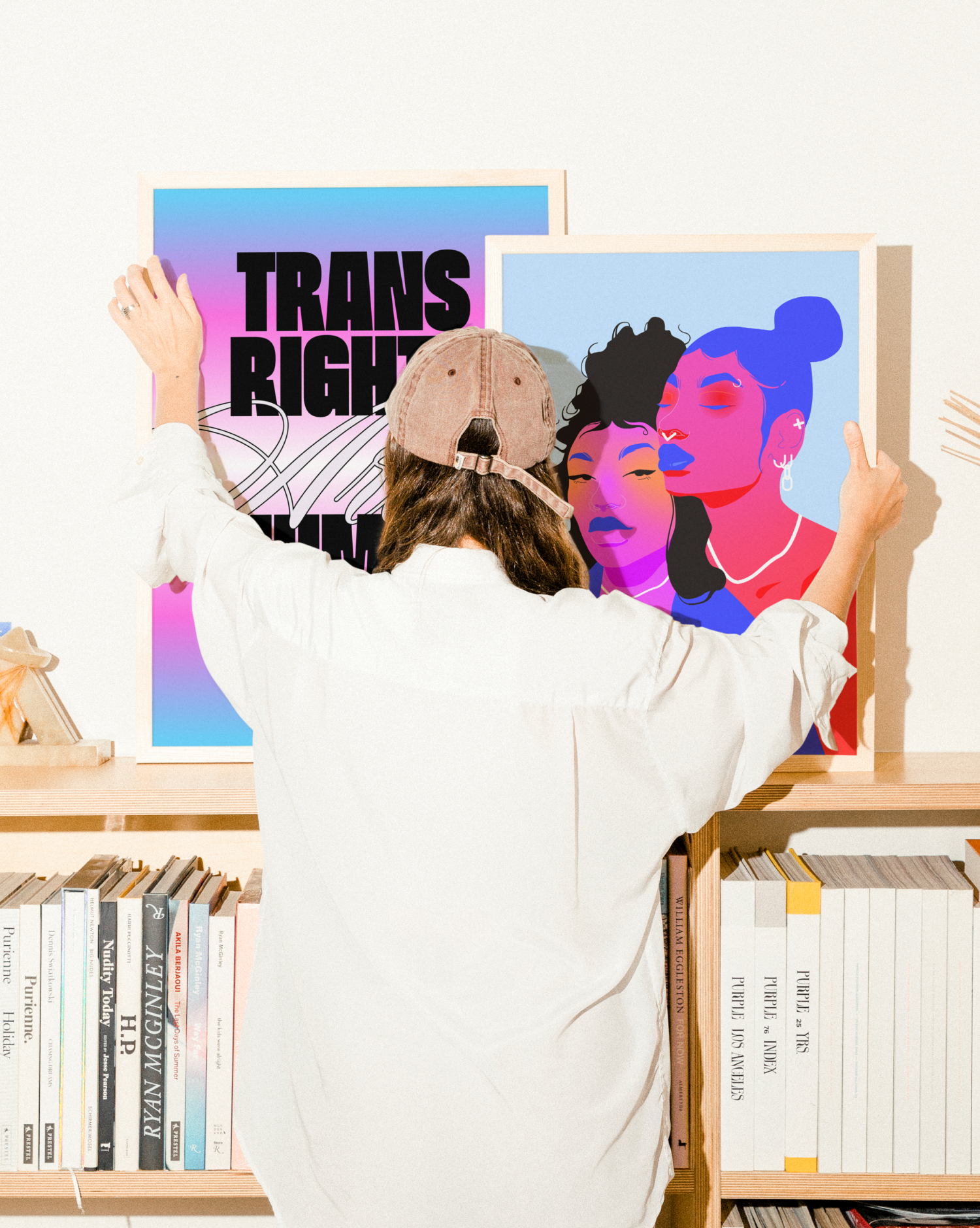 A person wearing a beige cap and white shirt is arranging two colorful posters on a bookshelf. The left poster has the text 'TRANS RIGHTS' in bold black letters on a pink and blue gradient background, and the right poster features a digital illustration of two women with their eyes closed, one with curly hair and the other with a bun, in vibrant pink and blue tones.