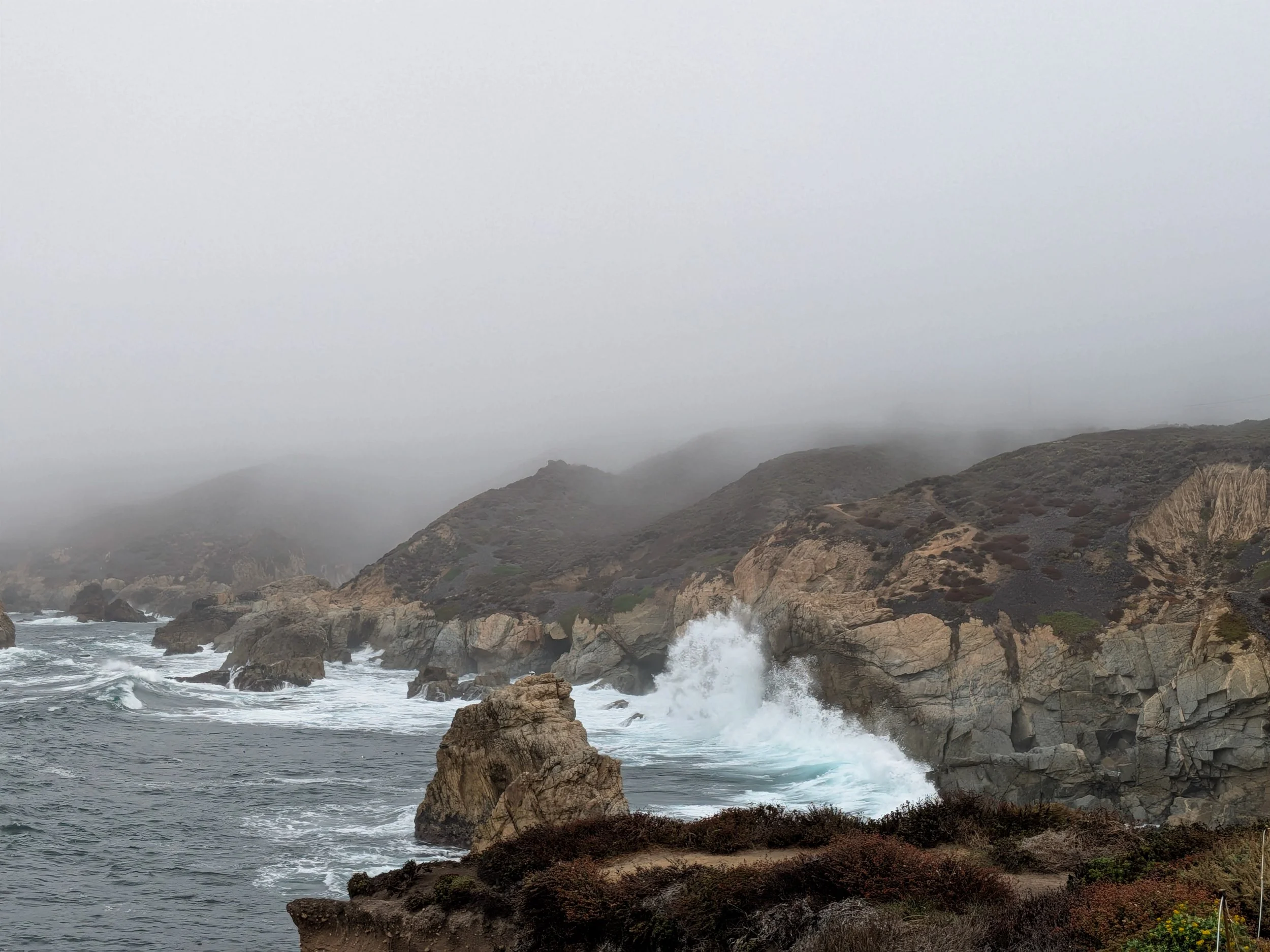 Coastal scene with waves crashing against rocky cliffs on a foggy day.