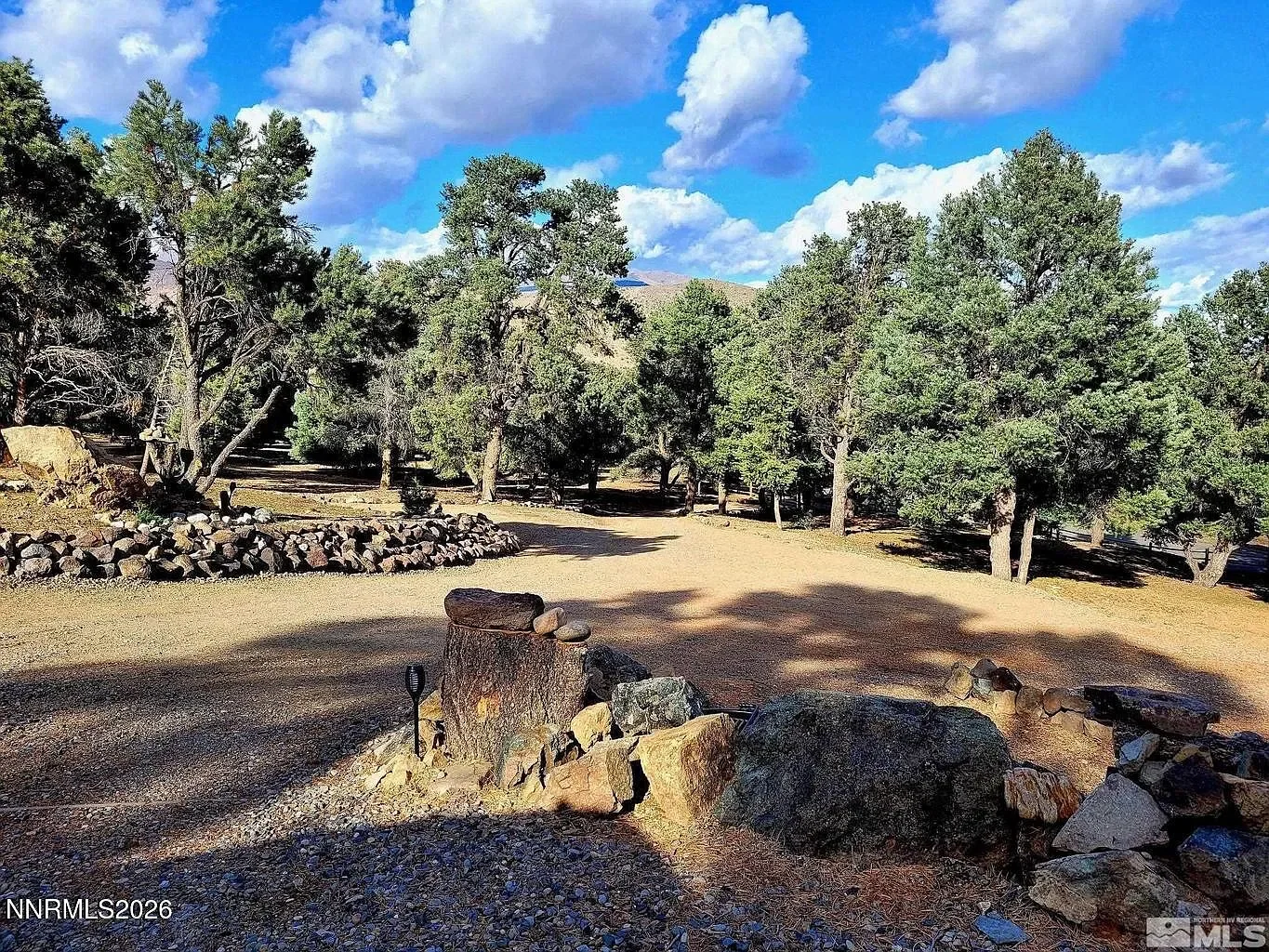 Scenic outdoor park scene with tall pine trees under a partly cloudy blue sky, surrounded by a circular arrangement of rocks and a tree stump in the foreground.
