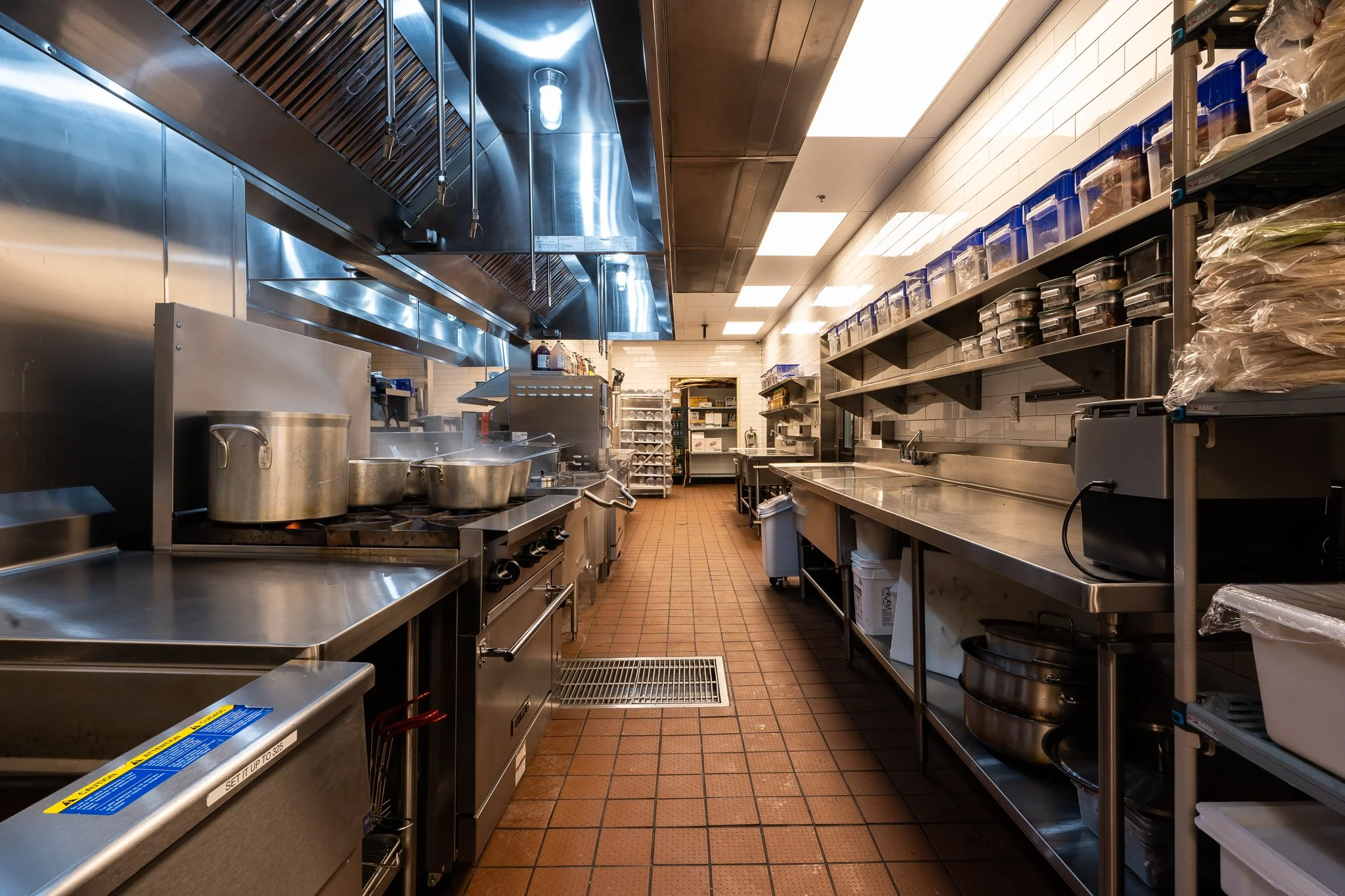 Commercial kitchen with stainless steel appliances and shelves, multiple large pots, and organized storage containers.