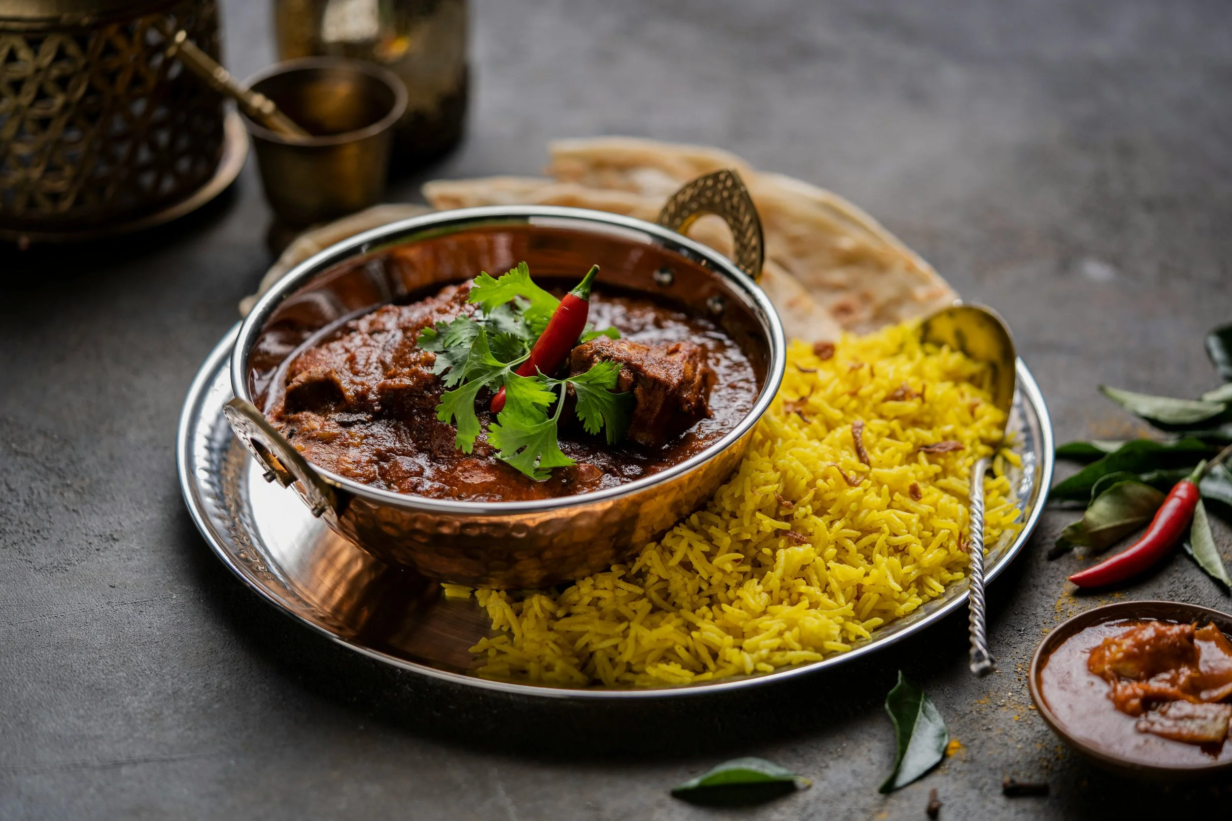 A traditional Indian meal with yellow rice, a bowl of beef curry garnished with cilantro and a red chili, and naan bread on a silver platter, with small bowls of sauce and chili peppers around.