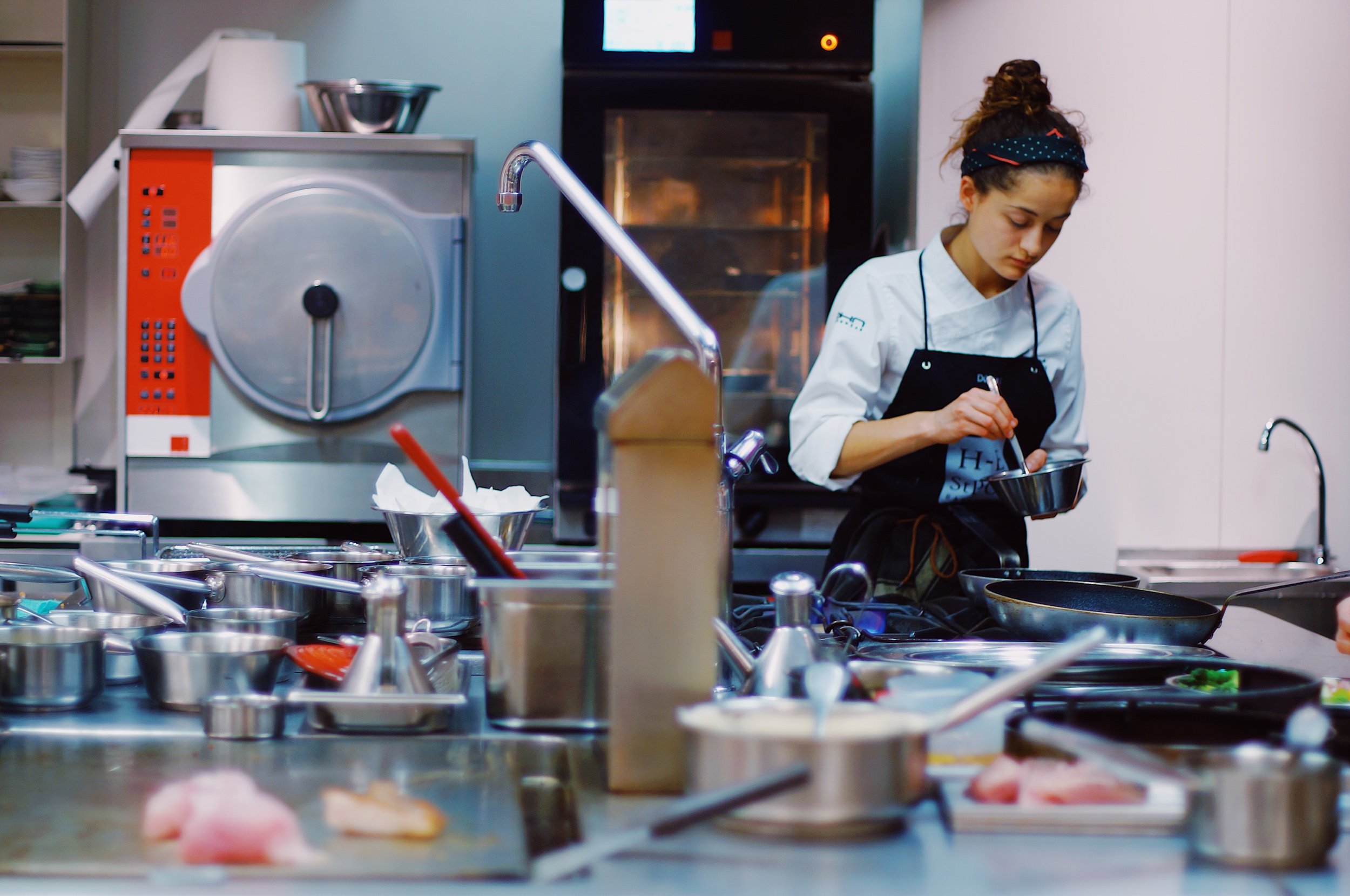 A young female chef in a professional kitchen preparing food with various pots, pans, and cooking utensils surrounding her.