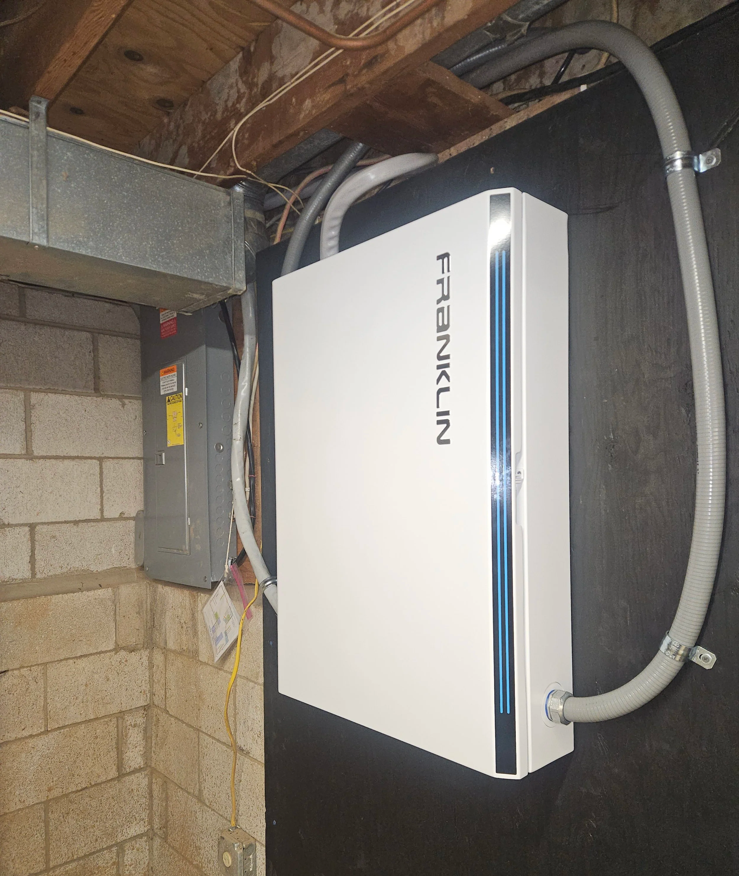 An electrical room with a Franklin whole home backup battery system mounted on a  wall, along with electrical wiring and a breaker box.