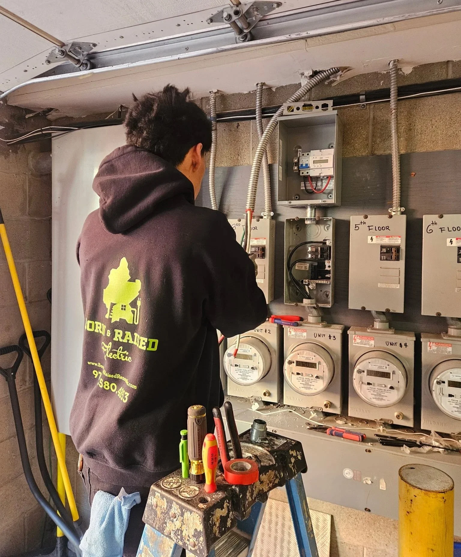 A licensed Born & Raised electrician providing electrical service on electrical meters and circuit breaker panels in a utility room surrounded by tools and wires.