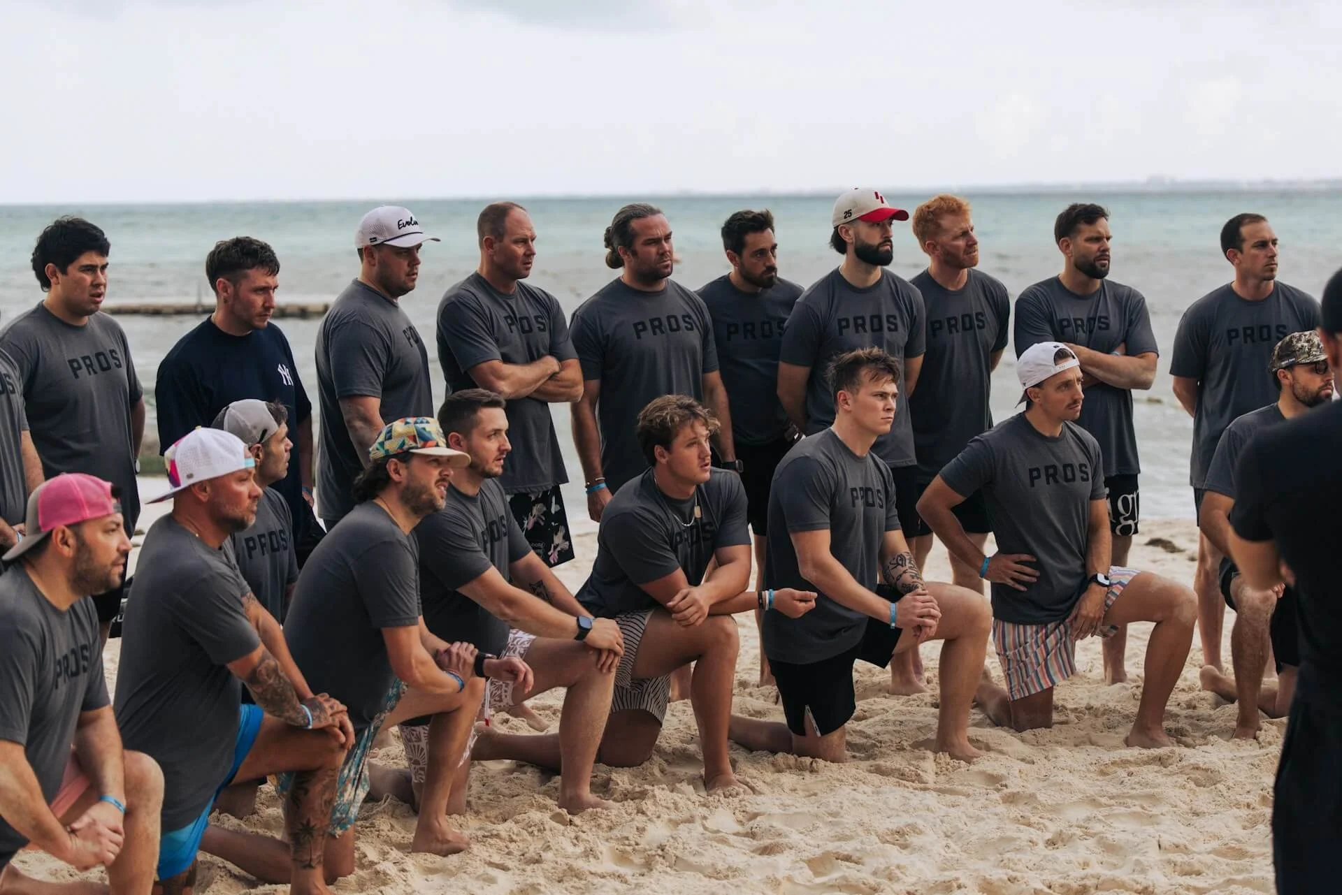 Group of men on a beach with kneeling positions, wearing gray T-shirts with 'PROS' printed on them, some in shorts, with a background of ocean water and cloudy sky.