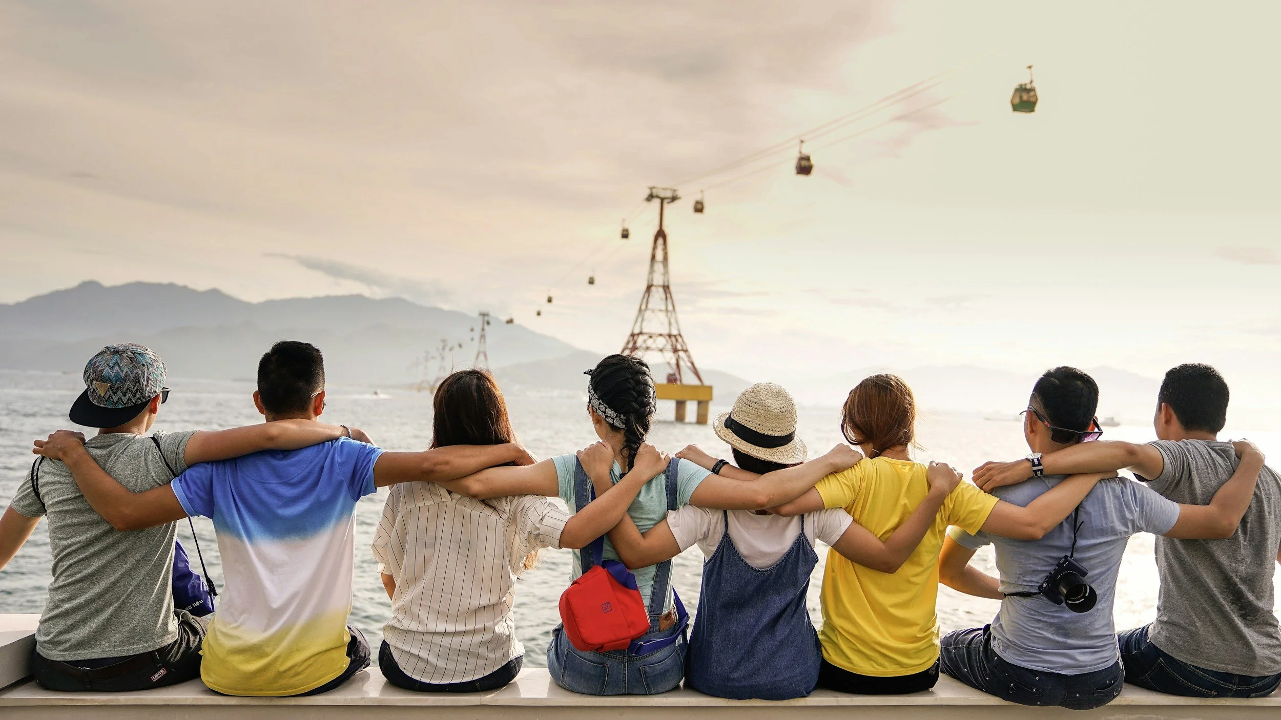 A diverse group of friends sitting on a dock with arms around each other, overlooking water and mountains while cable cars move across the sky.