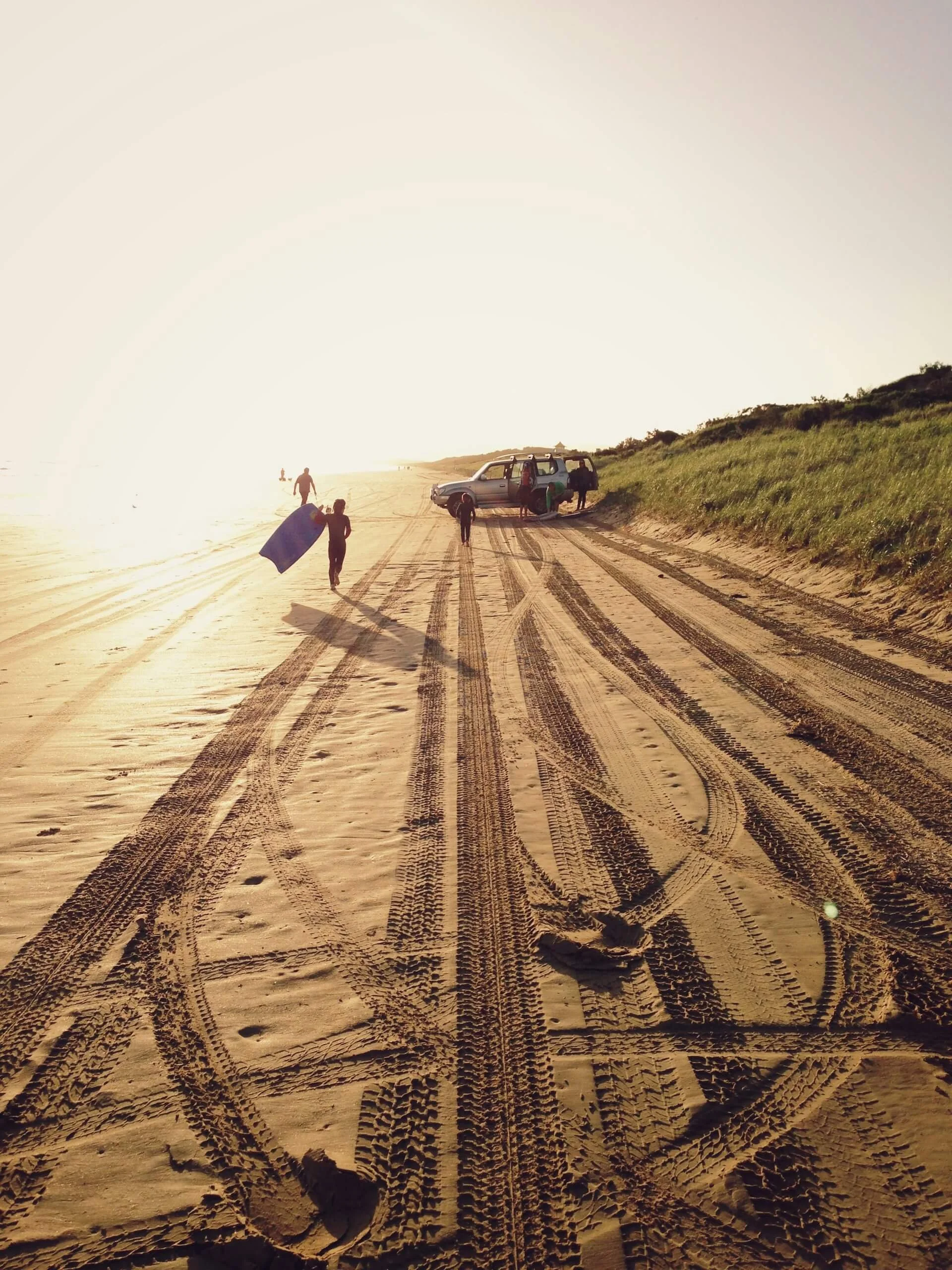 A beach with sand and tire tracks, with a silver vehicle and people near it, and green grass on a hill. The sun is setting or rising, casting a warm glow.