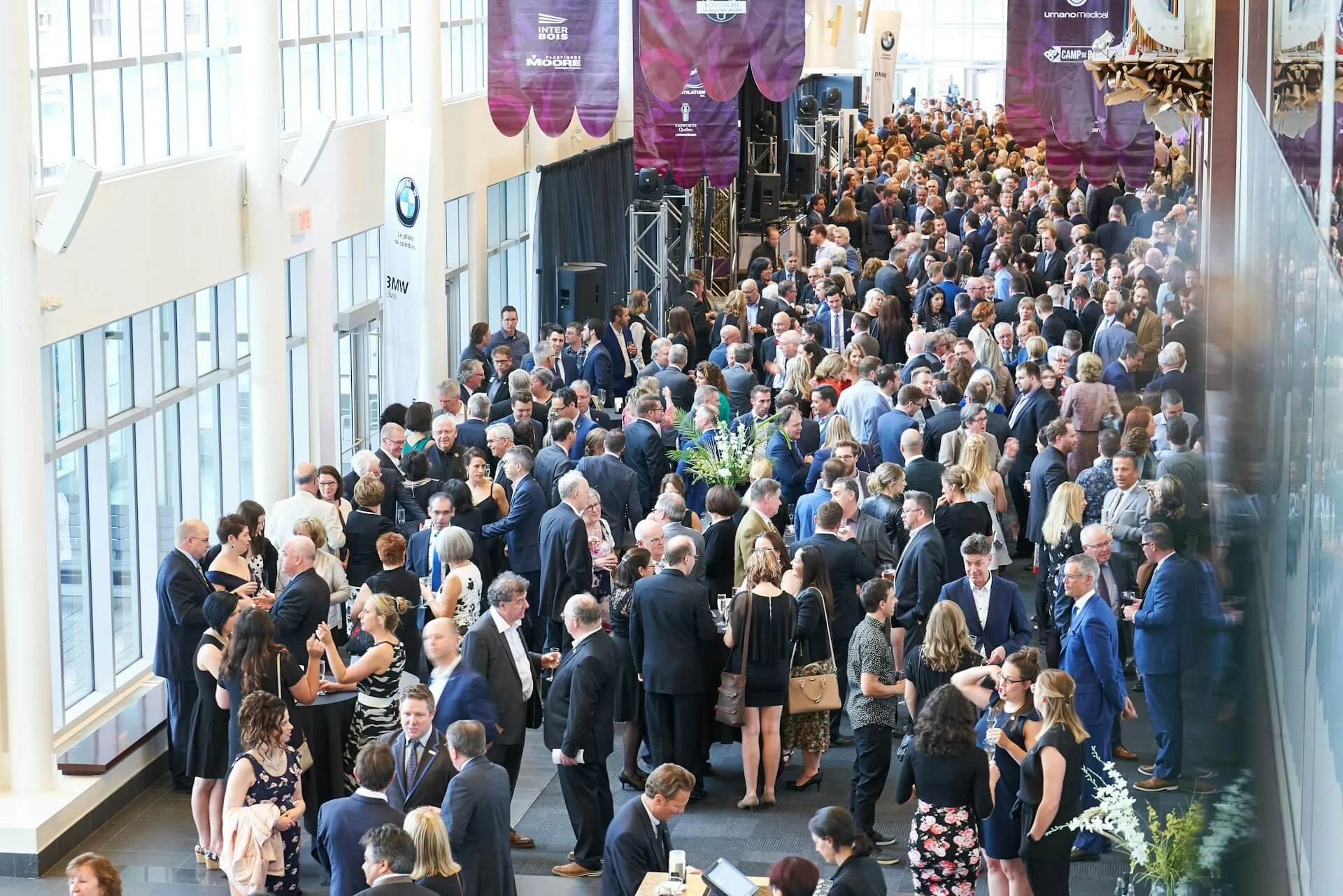 Large group of professionally dressed people socializing at a conference or networking event in a bright, modern venue with floor-to-ceiling windows and banners from companies like BMW and Umano Medical.