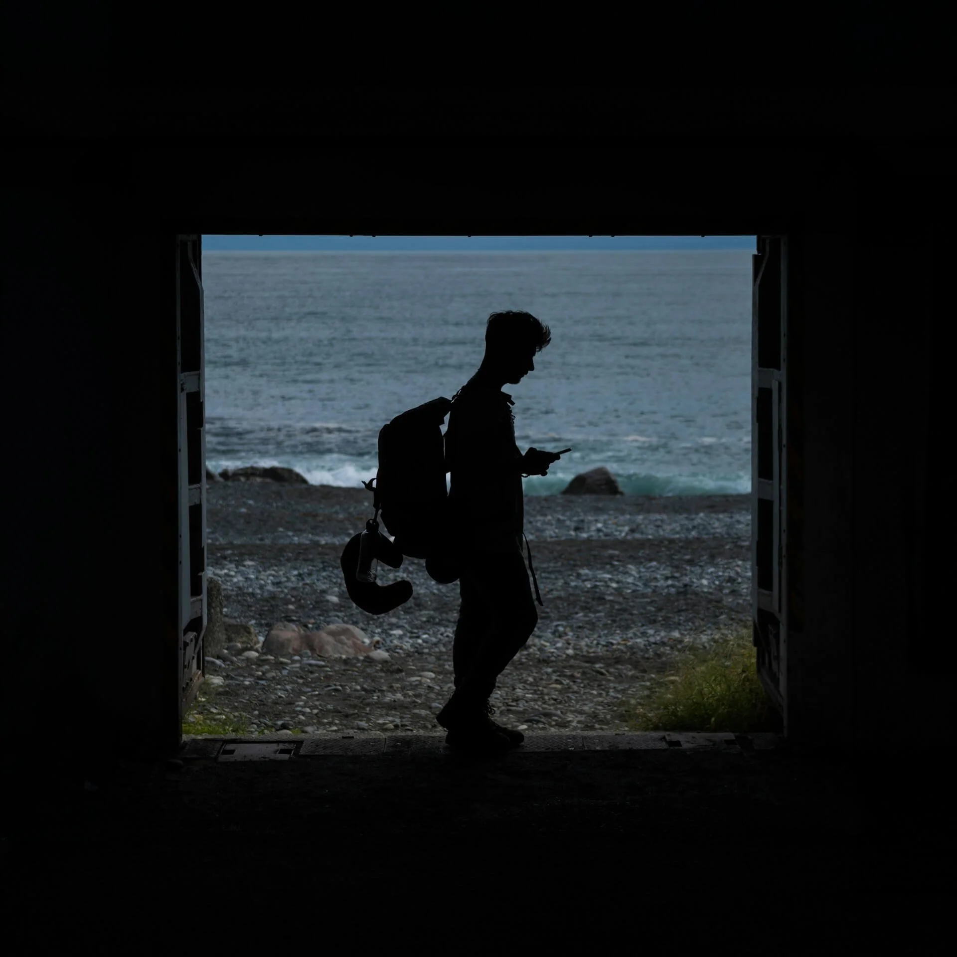 Person walking along a beach seen through a dark opening, with ocean in the background.