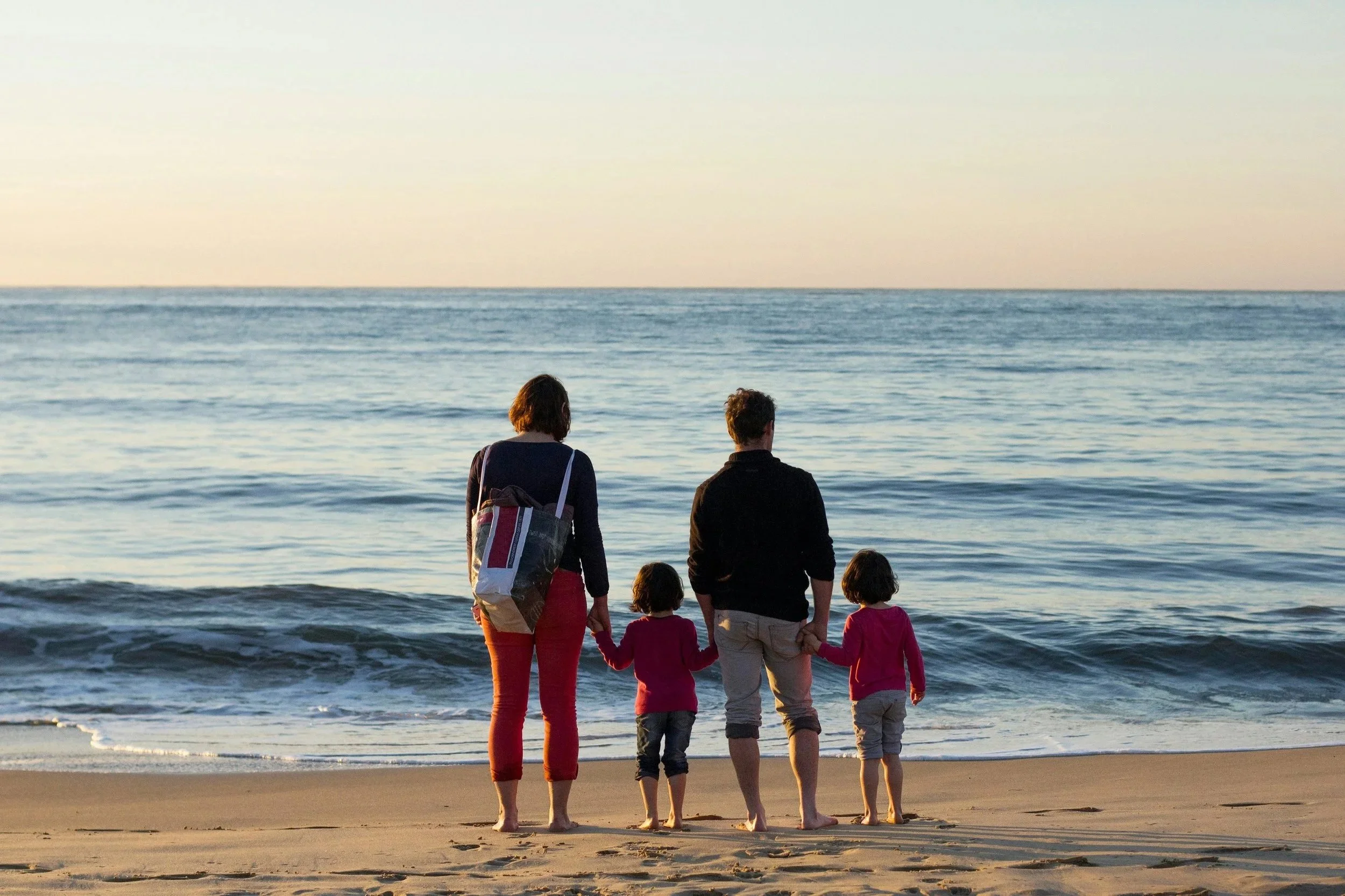 A family of five, including two adults and three children, standing at the edge of the ocean with waves, as the sun sets in the background.
