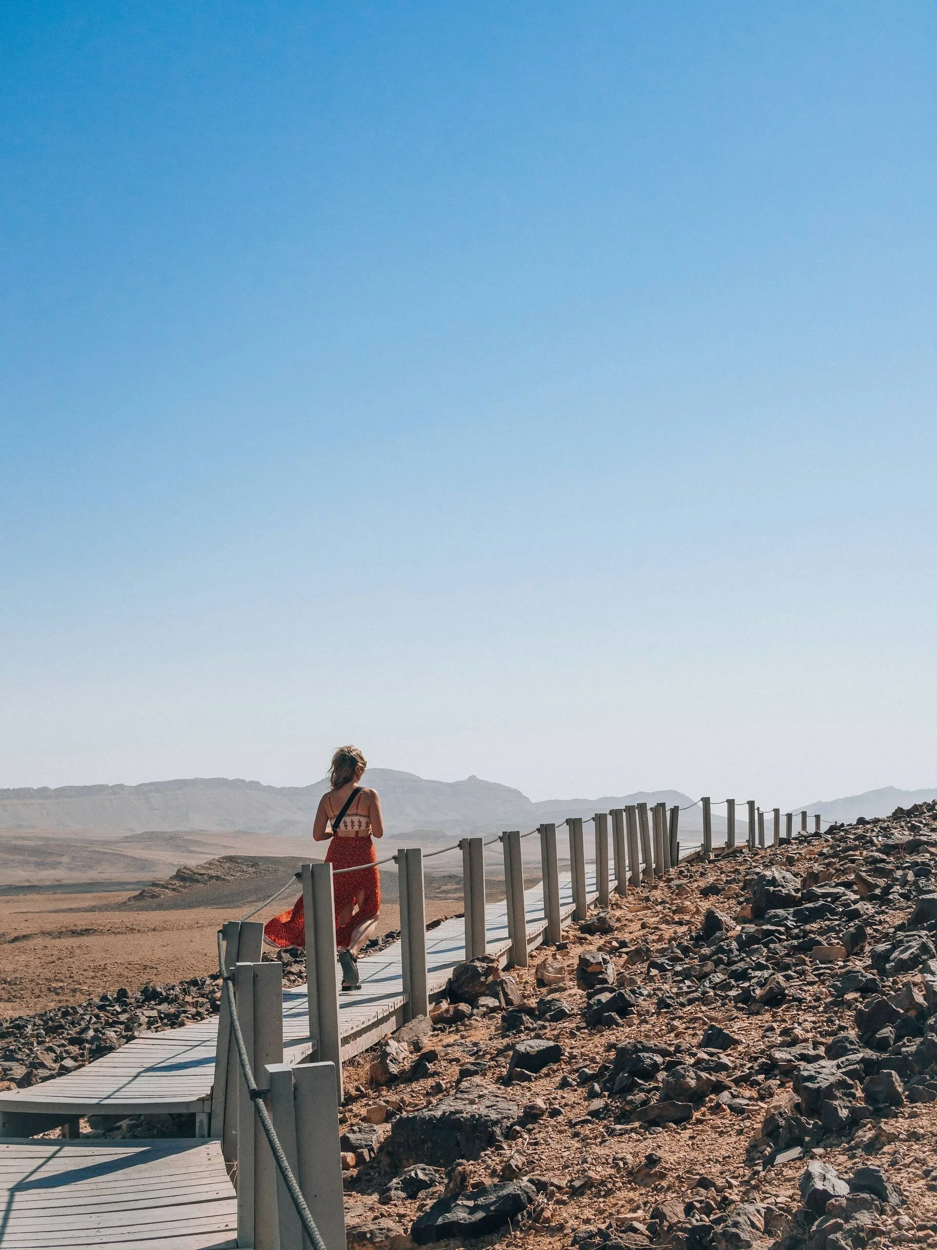 A woman in a red, patterned dress with a black shoulder bag walking on a wooden boardwalk through a rocky desert landscape with mountains in the distance and a clear blue sky.