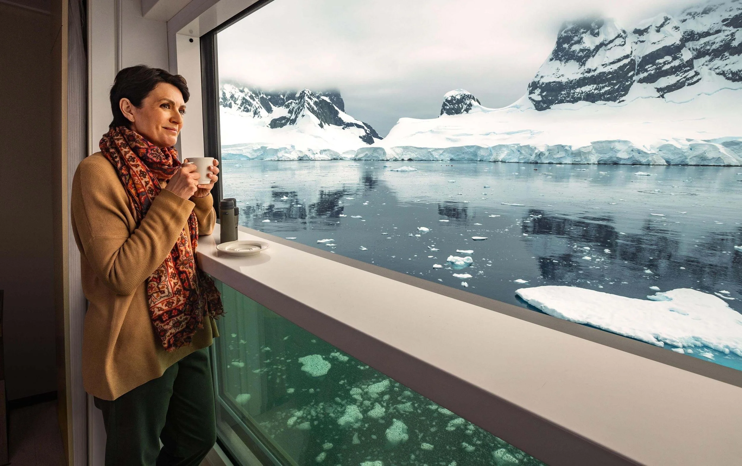 A woman holding a cup, looking out of a window at a glacier and ice-covered mountains in a cold, polar landscape.