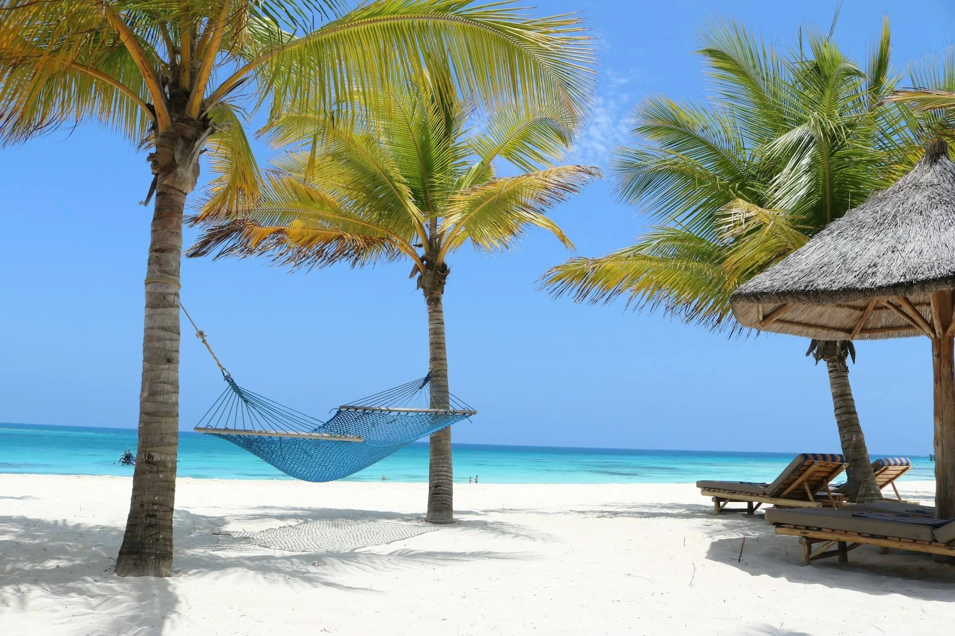 Tropical beach scene with palm trees, blue sky, hammock between two trees, and lounge chairs under a thatched umbrella on white sand.