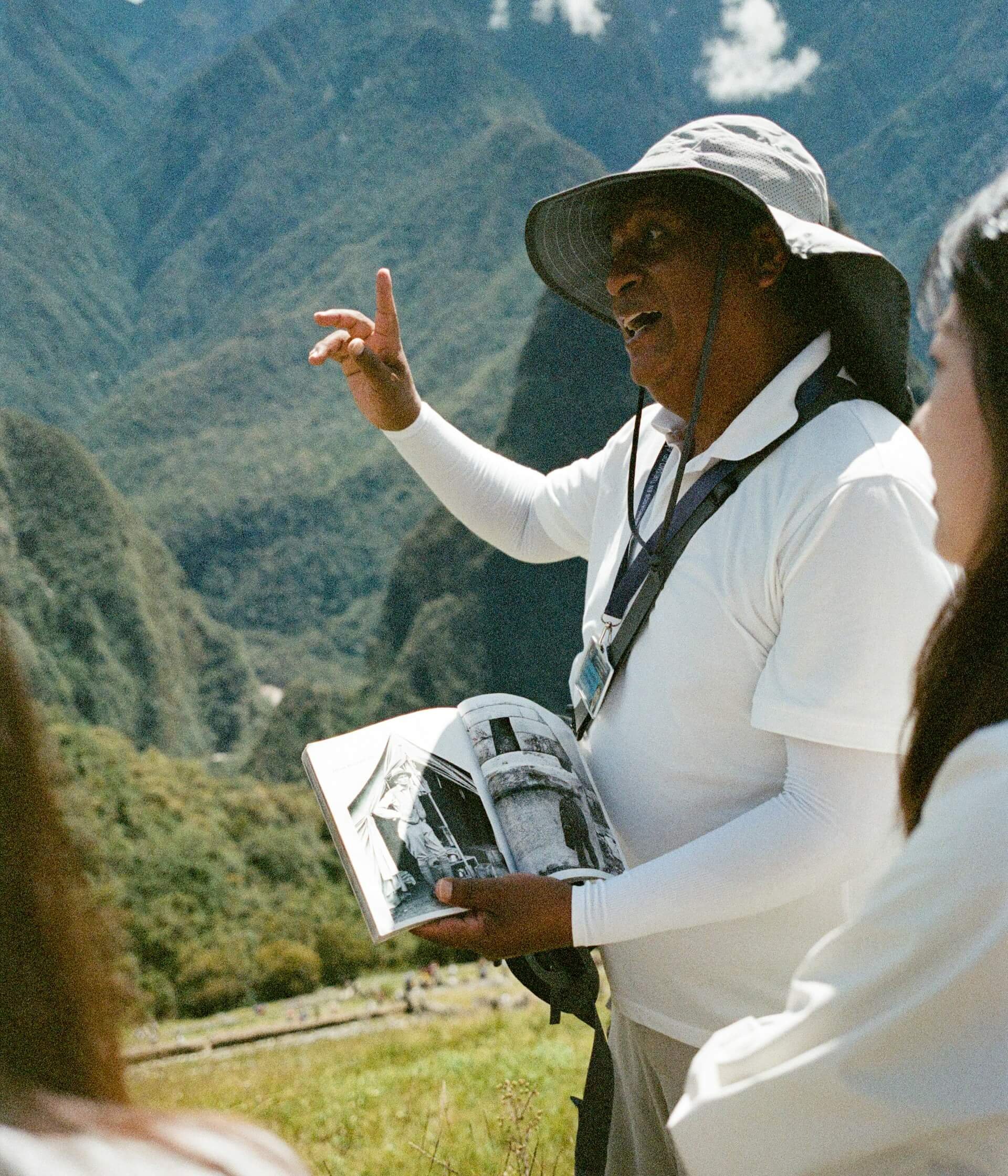 Person wearing a wide-brimmed hat, white shirt, and sunglasses, holding an open booklet, possibly reading or showing photographs, outdoors in a landscape with mountains and trees.