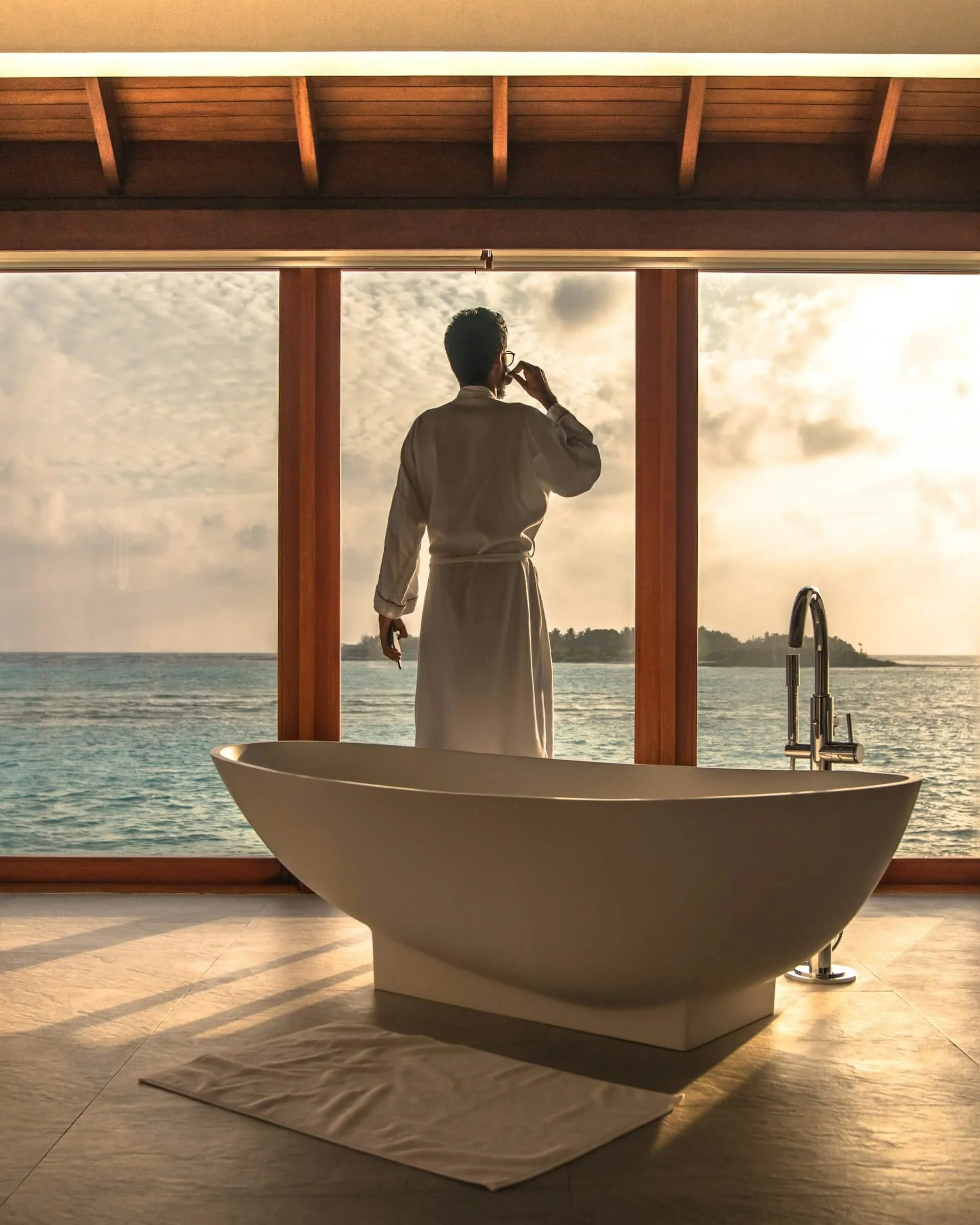 A person dressed in a white robe and wearing glasses is standing near a large window overlooking a body of water and a cloudy sky at sunset. The person appears to be thinking or meditating. A modern freestanding bathtub is visible in the foreground inside a room with wooden floors.